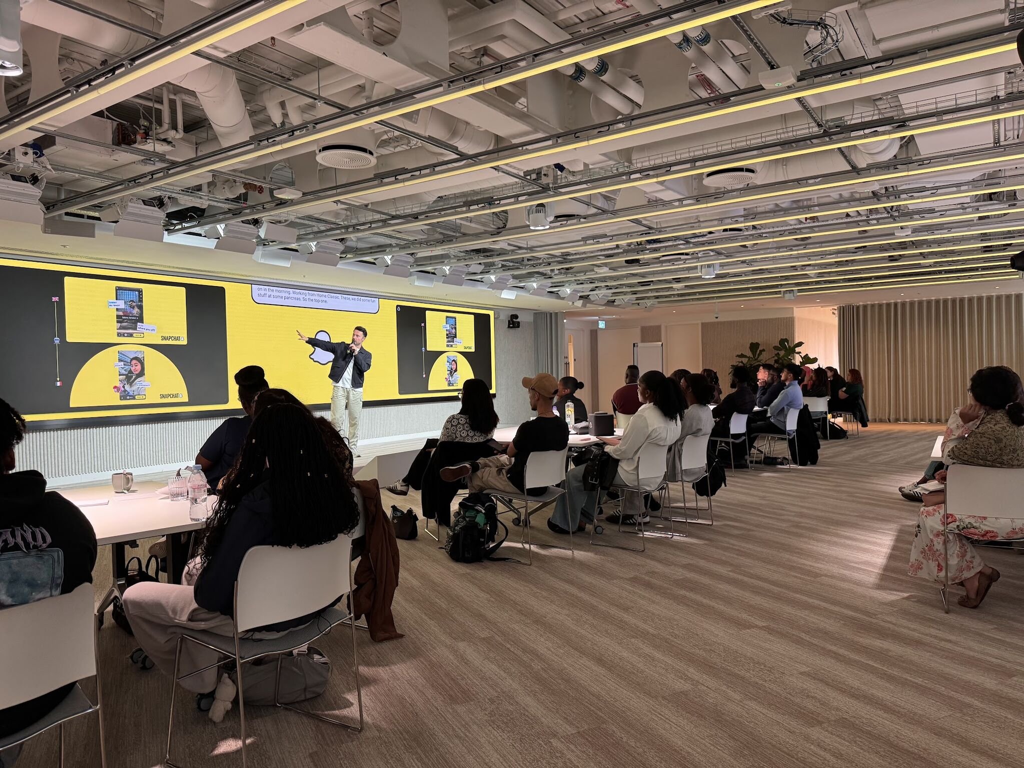 A conference room with a man giving a presentation to an audience seated at white tables. Large yellow digital screens display content related to Snapchat, and the ceiling has exposed pipes and lighting.