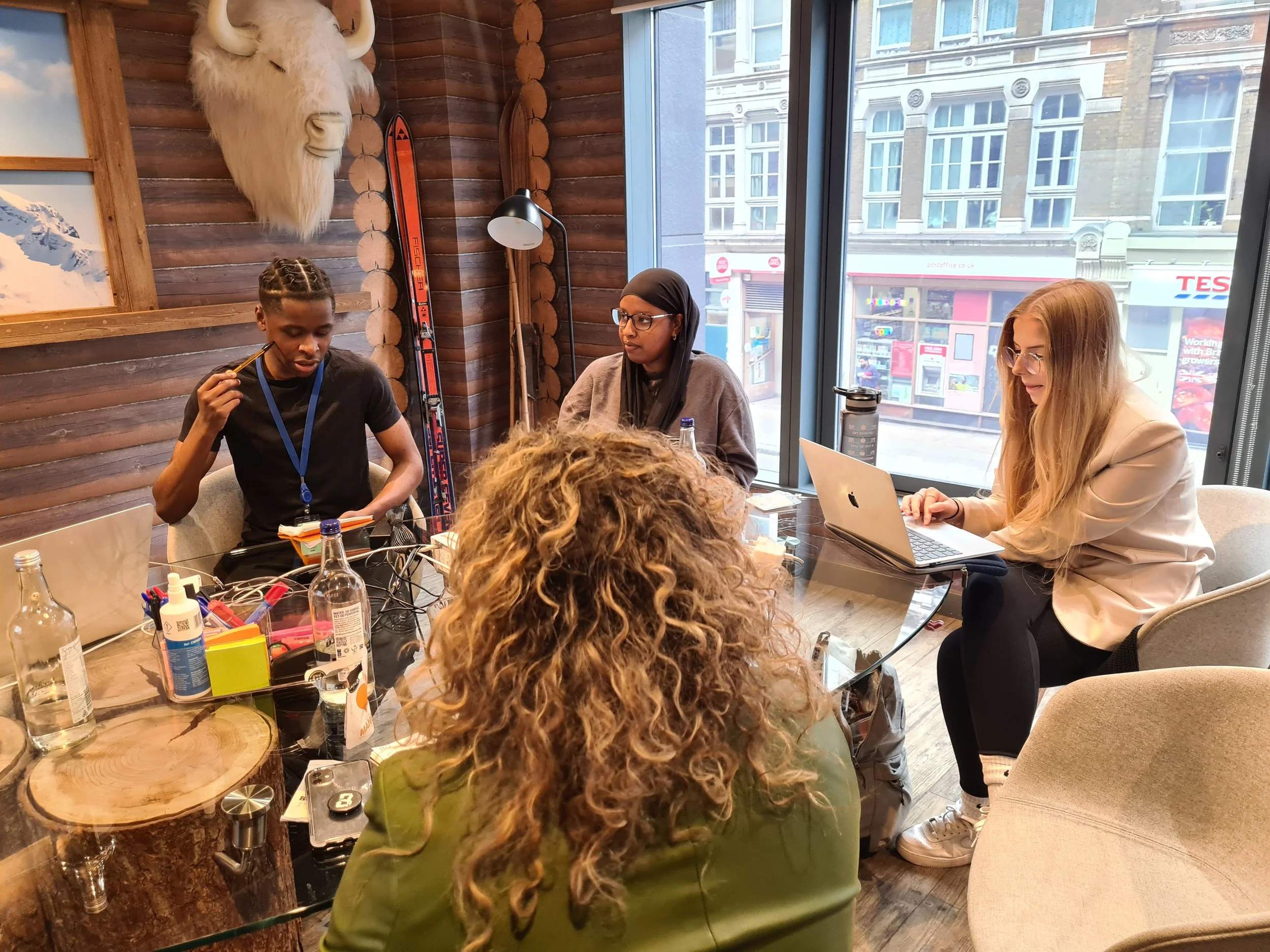 A group of people sitting around a table in a cozy, wood-paneled room with a large window showing buildings outside. The table has various bottles, papers, and electronic devices. A woman with long blonde hair and glasses is working on a laptop, and a young man with braided hair is holding a pencil. A woman wearing a hijab and glasses is also present. The wall decor includes a mounted mountain goat head and skis.
