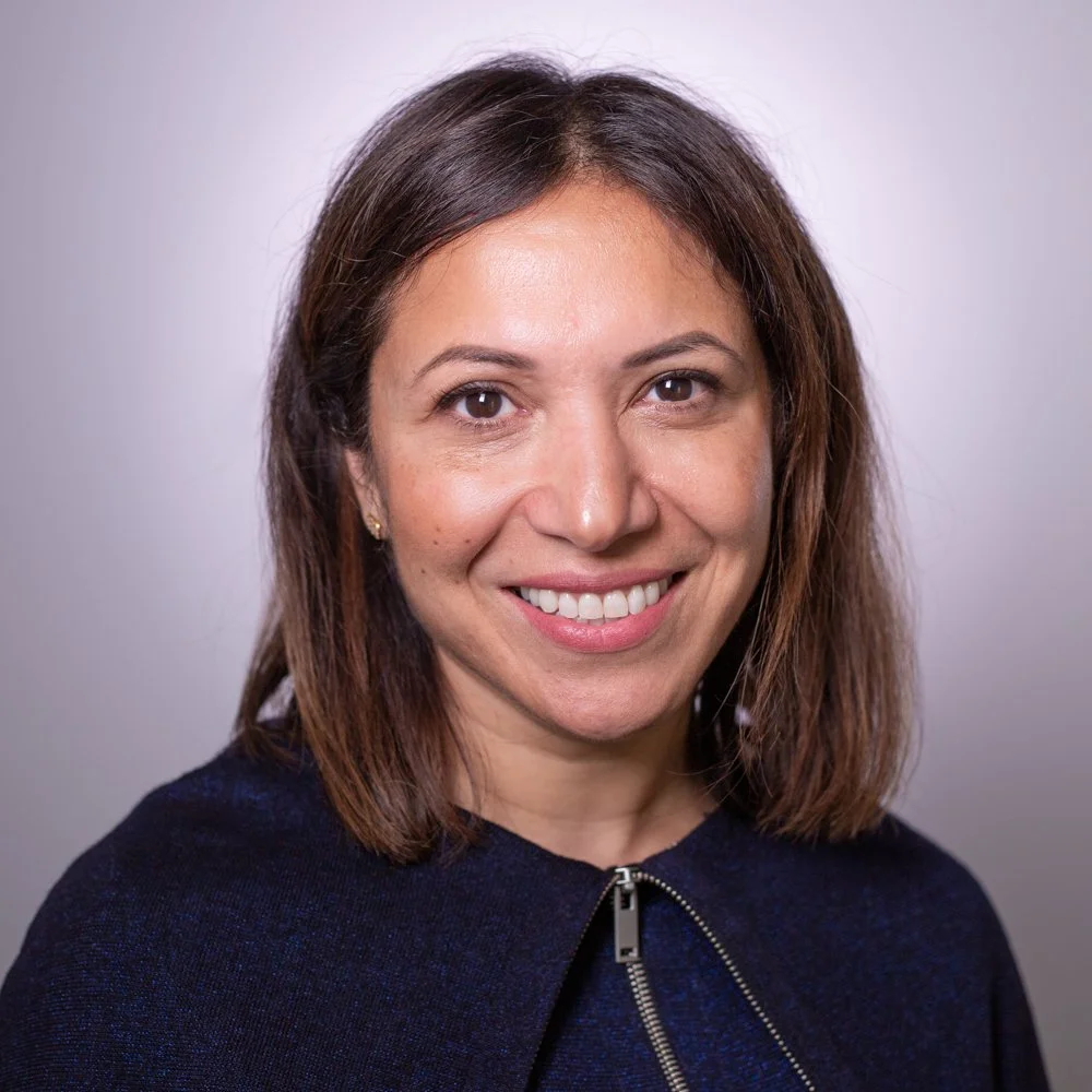 A portrait of a smiling woman with shoulder-length brown hair, wearing a dark navy top, against a plain light gray background.