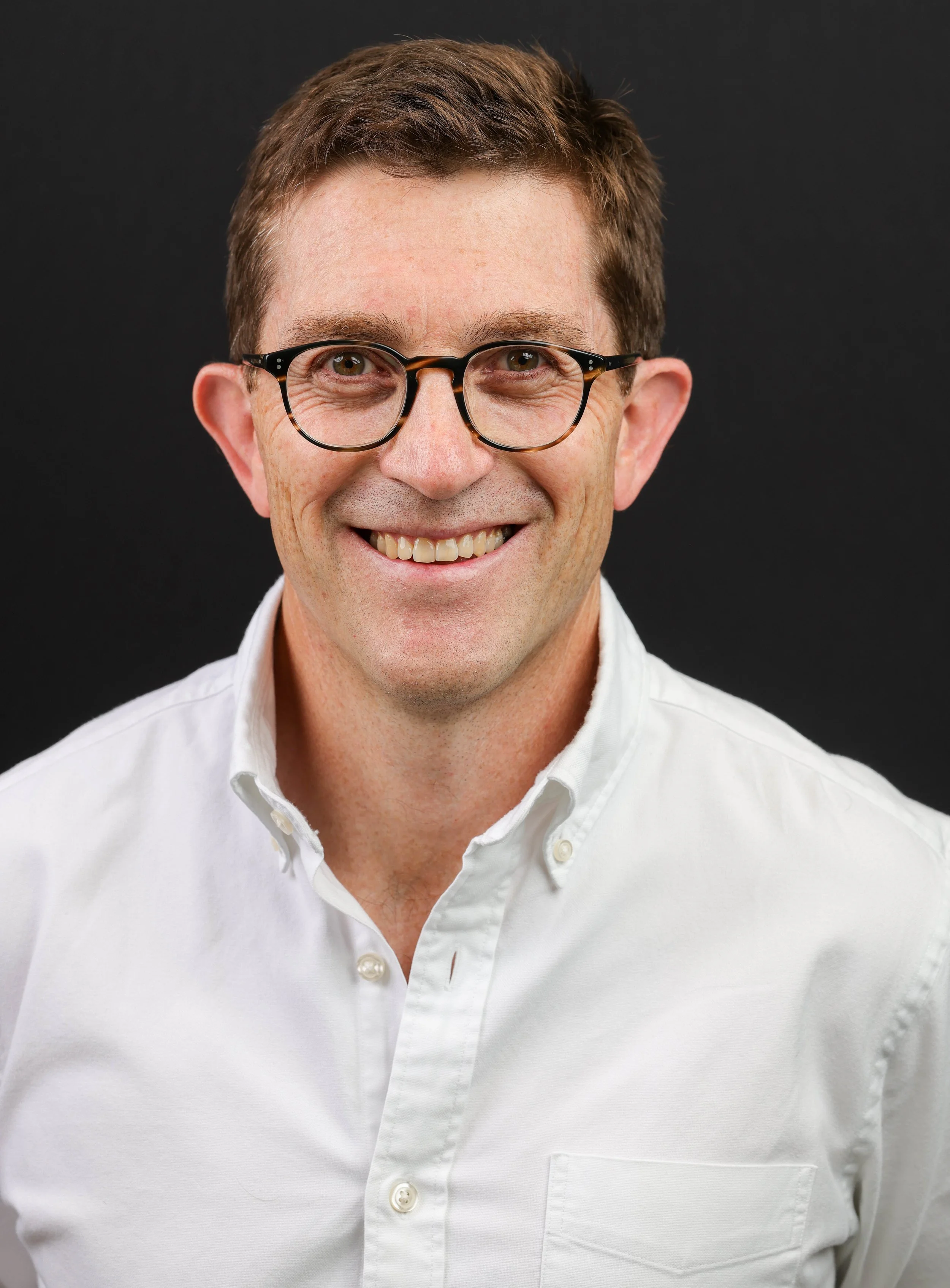 Close-up of a smiling man wearing glasses and a white shirt against a black background.