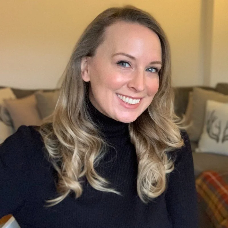 A woman with blonde, wavy hair, wearing a black turtleneck sweater, smiling in a cozy living room with beige walls and throw pillows on a couch.