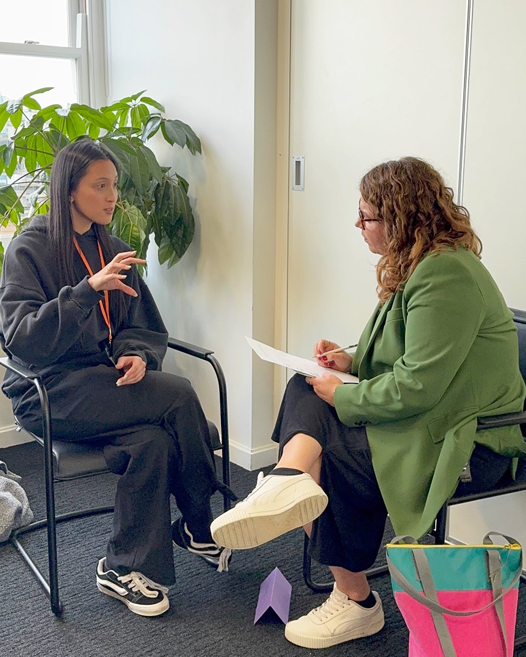 Two women are engaged in a conversation in a room. One woman with long dark hair, dressed in a black hoodie and pants, is sitting on a black chair and gesturing with her hand. The other woman with curly red hair, wearing glasses, a green blazer, and black pants, is sitting on another black chair and taking notes on a notepad. There is a large green plant and a window in the background.
