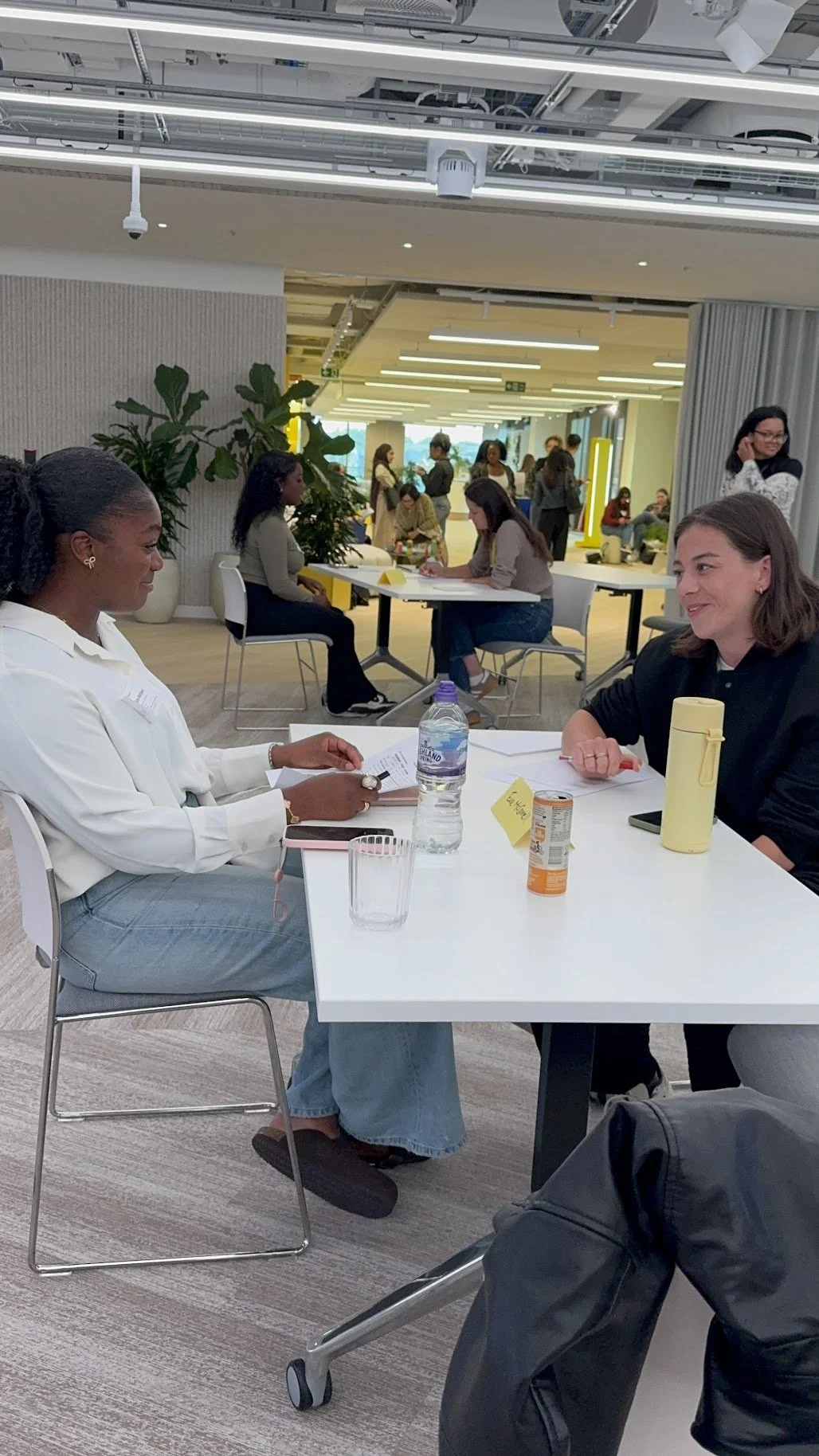 Two women sitting at a table engaged in conversation in a modern office or conference space, with several other people working and socializing in the background.