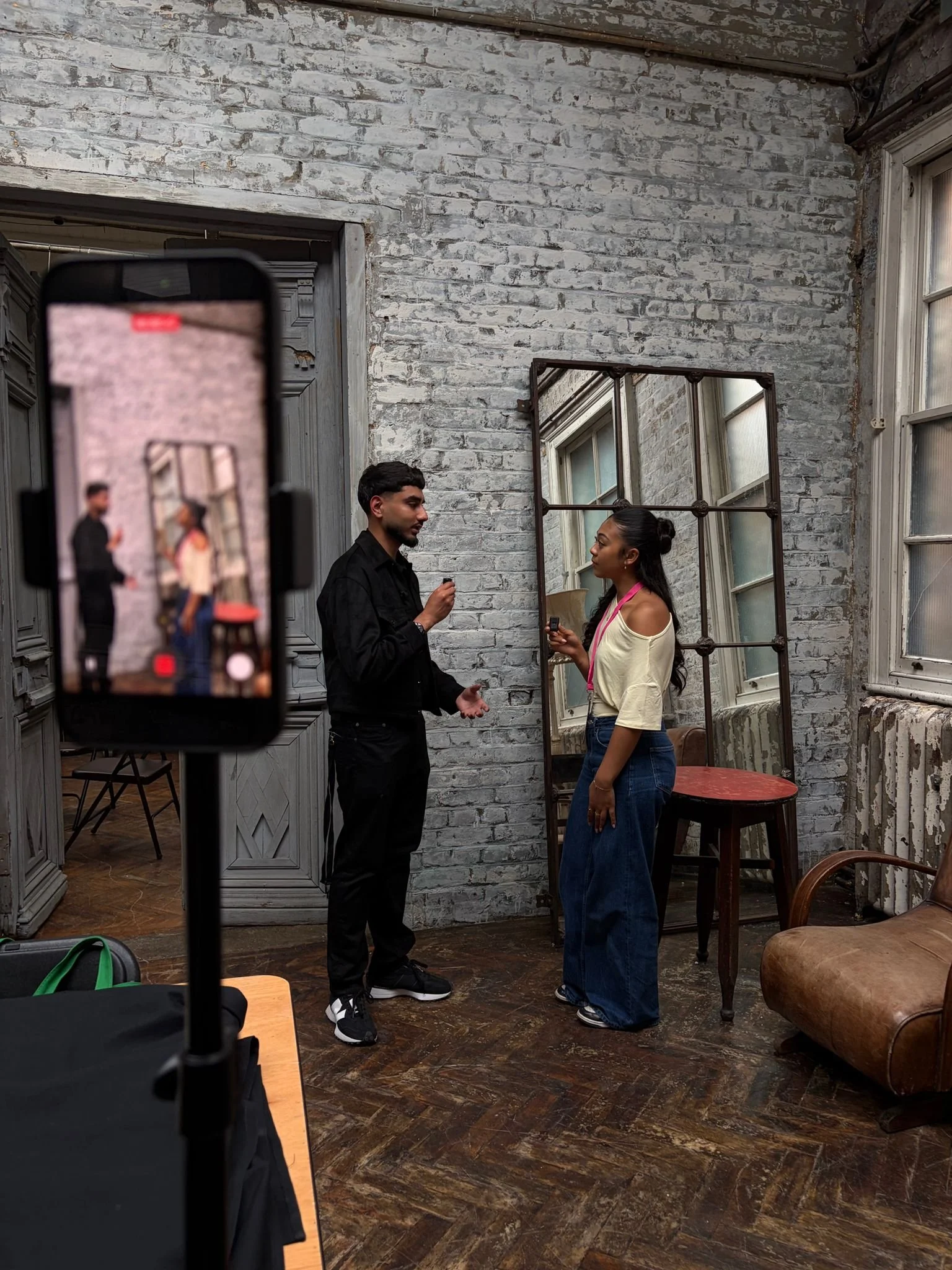 A man and woman engaging in a conversation in an industrial-style room with white brick walls and large windows. The man is holding an object, and the woman is listening. A mirror is behind them, reflecting parts of the room. A smartphone on a tripod is recording or taking a picture of them.
