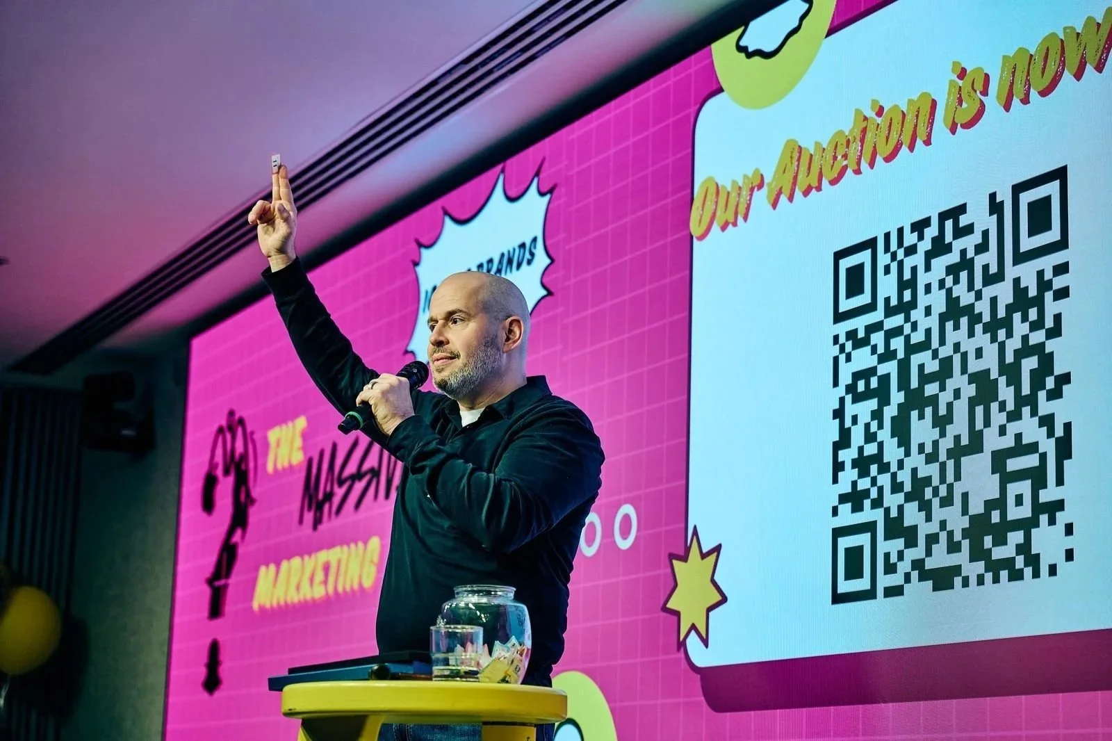 A man with a beard speaking into a microphone during a presentation, with a colorful slide projected behind him that includes a QR code and marketing related text.