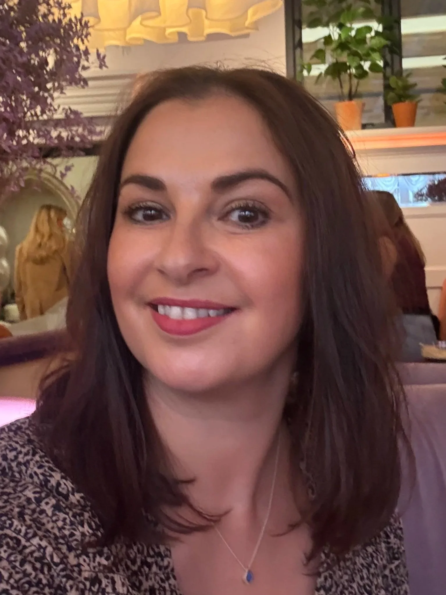 A woman with shoulder-length brown hair smiling, wearing a patterned top and a blue pendant necklace, in a warmly lit restaurant or cafe with decorative plants and wall art in the background.