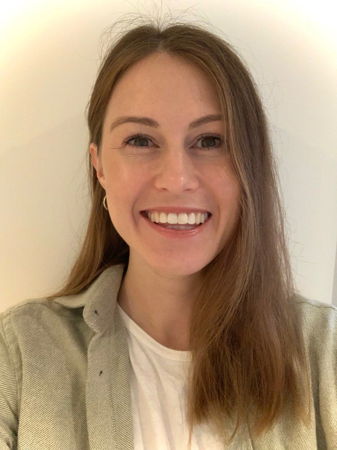 A woman with long reddish-brown hair smiling while taking a selfie against a plain white wall.