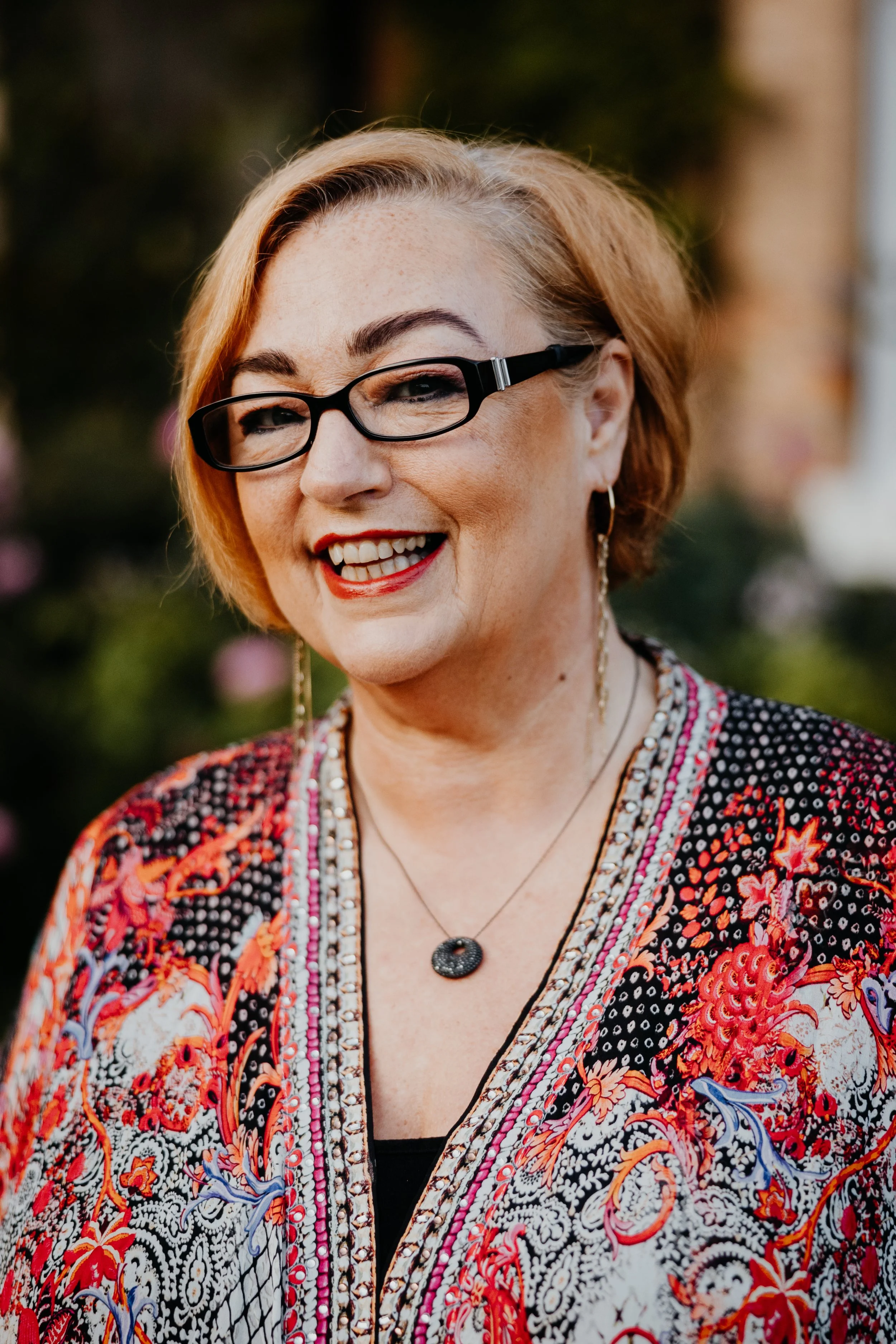 Close-up photo of a smiling woman with short red hair wearing glasses, earrings, and a colorful patterned blouse with floral and geometric designs.
