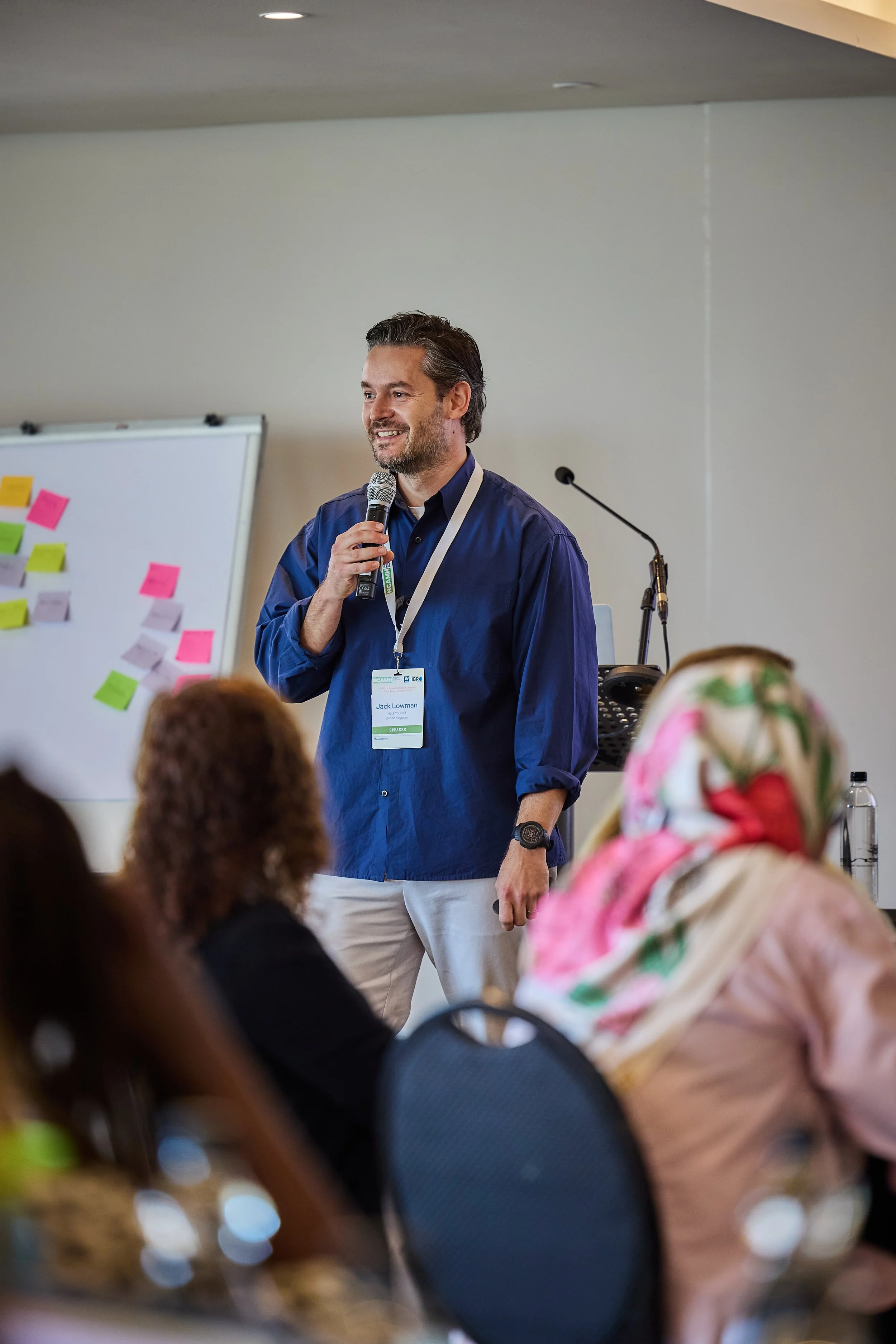 A man with dark hair and a beard speaking into a microphone, wearing a blue shirt and a conference badge, standing in front of a whiteboard with colorful sticky notes. Seated audience, some women wearing headscarves, are facing him in a conference ro