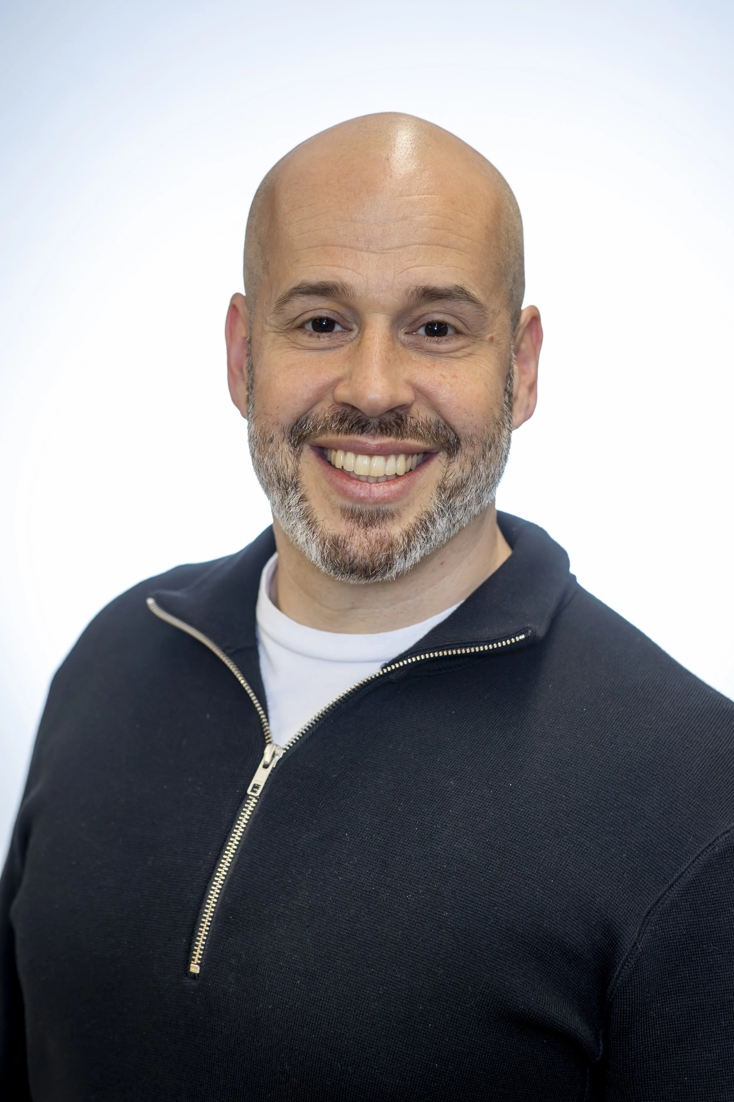 A man with a bald head and a beard, smiling, wearing a black jacket with a zip, against a plain light background.