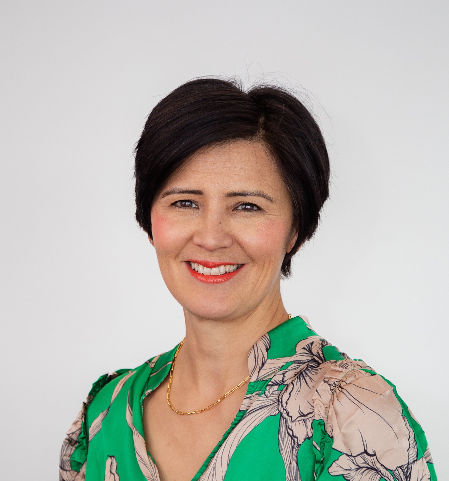 Portrait of a woman with short black hair, smiling, wearing a green floral dress and a gold necklace, against a plain light gray background.