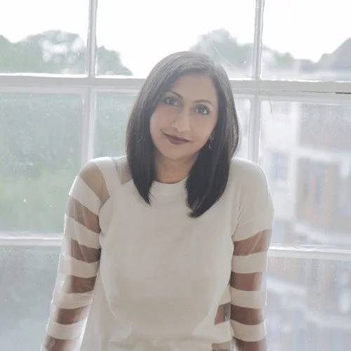 A woman with shoulder-length dark hair, wearing a white top with sheer striped sleeves, standing in front of a large window with a view of buildings outside.