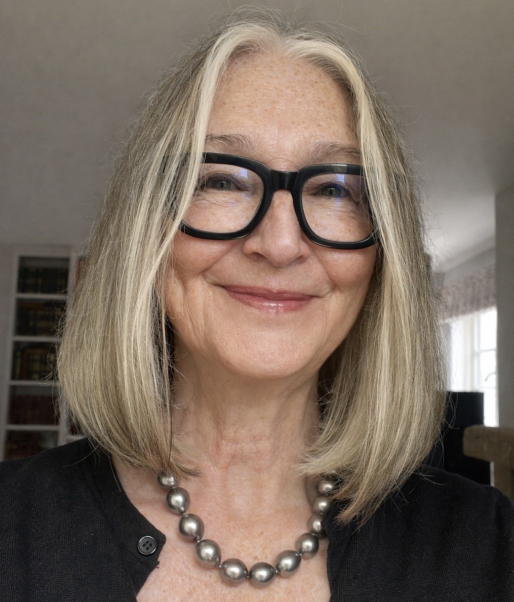 A woman with shoulder-length blonde hair, wearing black glasses, a pearl necklace, and a black top, smiling indoors with bookshelves and a window in the background.