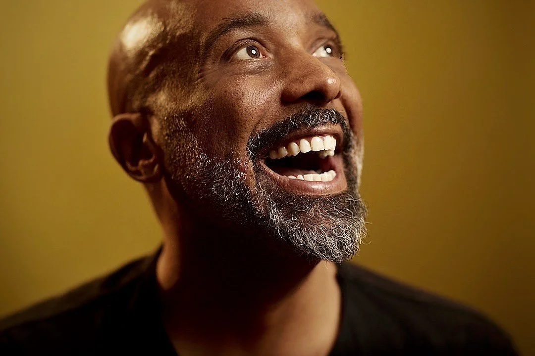 A joyful middle-aged man with a beard and mustache, smiling and looking upward, wearing a black shirt against a yellow background.