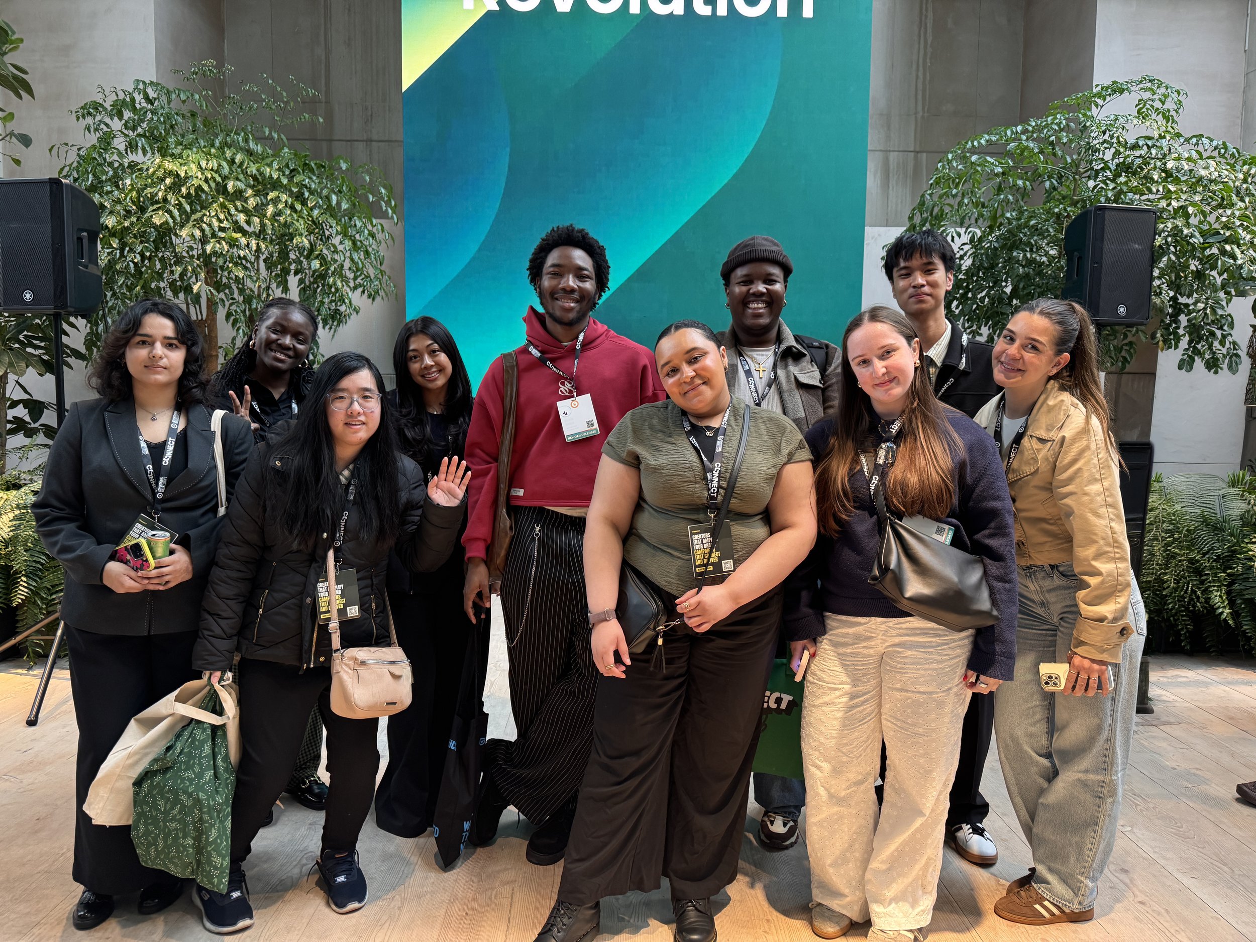 A diverse group of eleven young adults posing for a photo at an indoor event. They are standing in front of a large green and teal banner with partial word 'Revolution' visible. The group is smiling and making various gestures, with some wearing conference lanyards and badges. The background includes large green plants and a concrete wall.