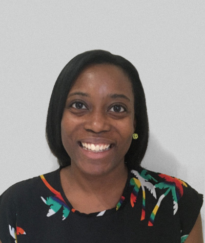 A woman smiling at the camera, wearing a black top with colorful floral patterns and a green smiley face earring, standing against a plain white background.