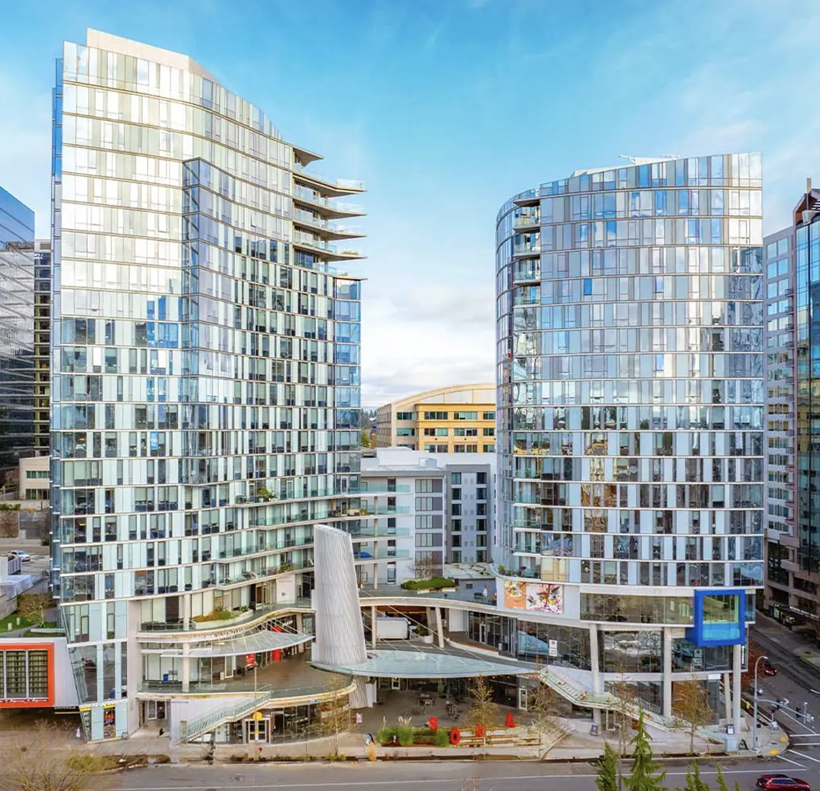 Two modern glass high-rise buildings with curved facades and balconies, part of a shopping mall or commercial complex, located in an urban area with other buildings and a street in the foreground.