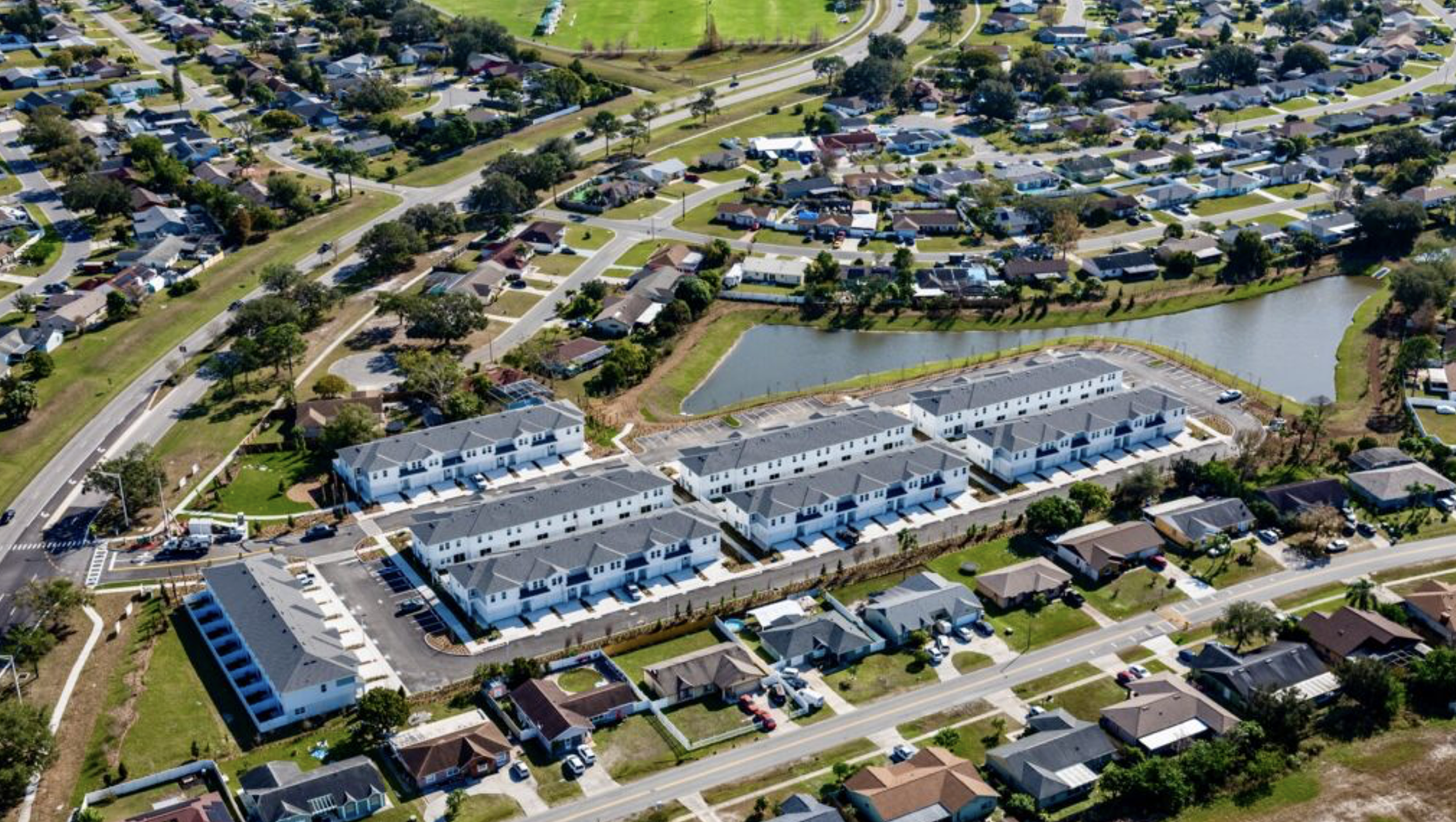 An aerial view of a residential neighborhood with a pond, a cluster of white townhouses, and surrounding single-family homes with neatly maintained lawns and roads.