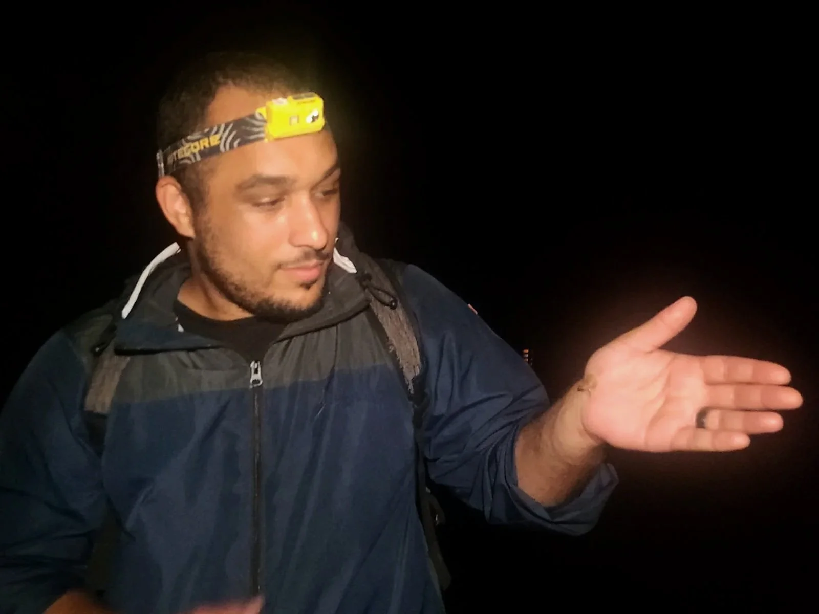 A man wearing a headlamp and a dark jacket is standing in a dark environment, gesturing with his right hand as if explaining something.