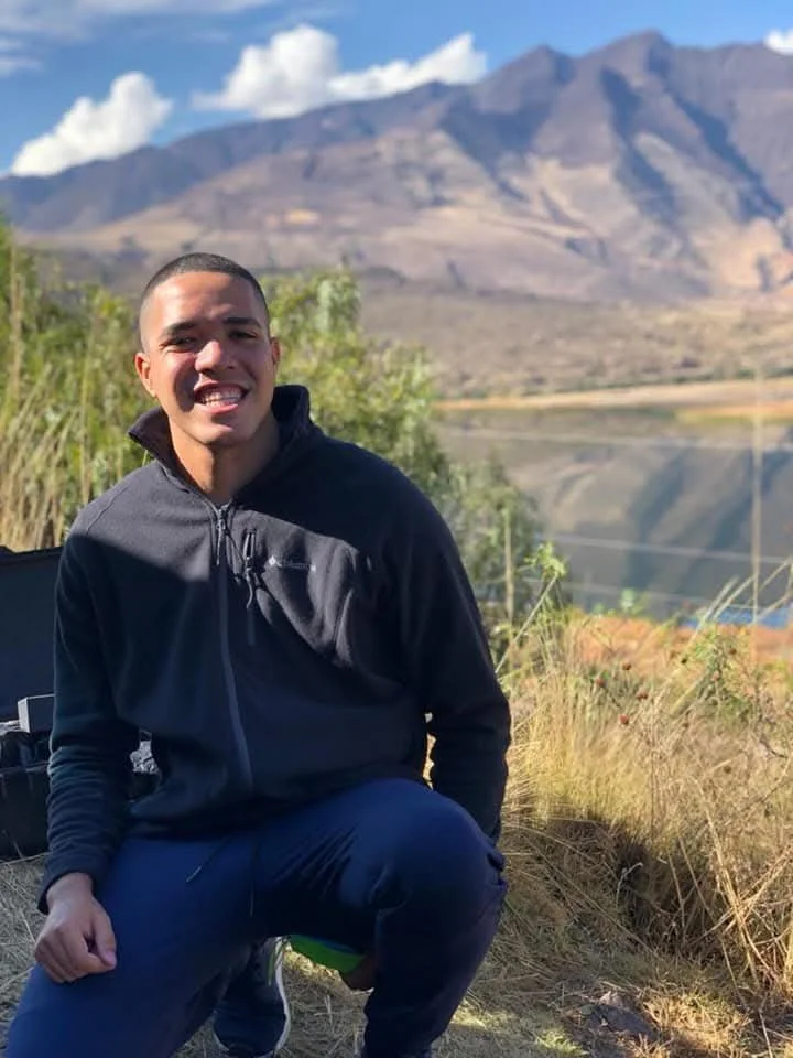 A man smiling outdoors near a lake with mountains in the background.
