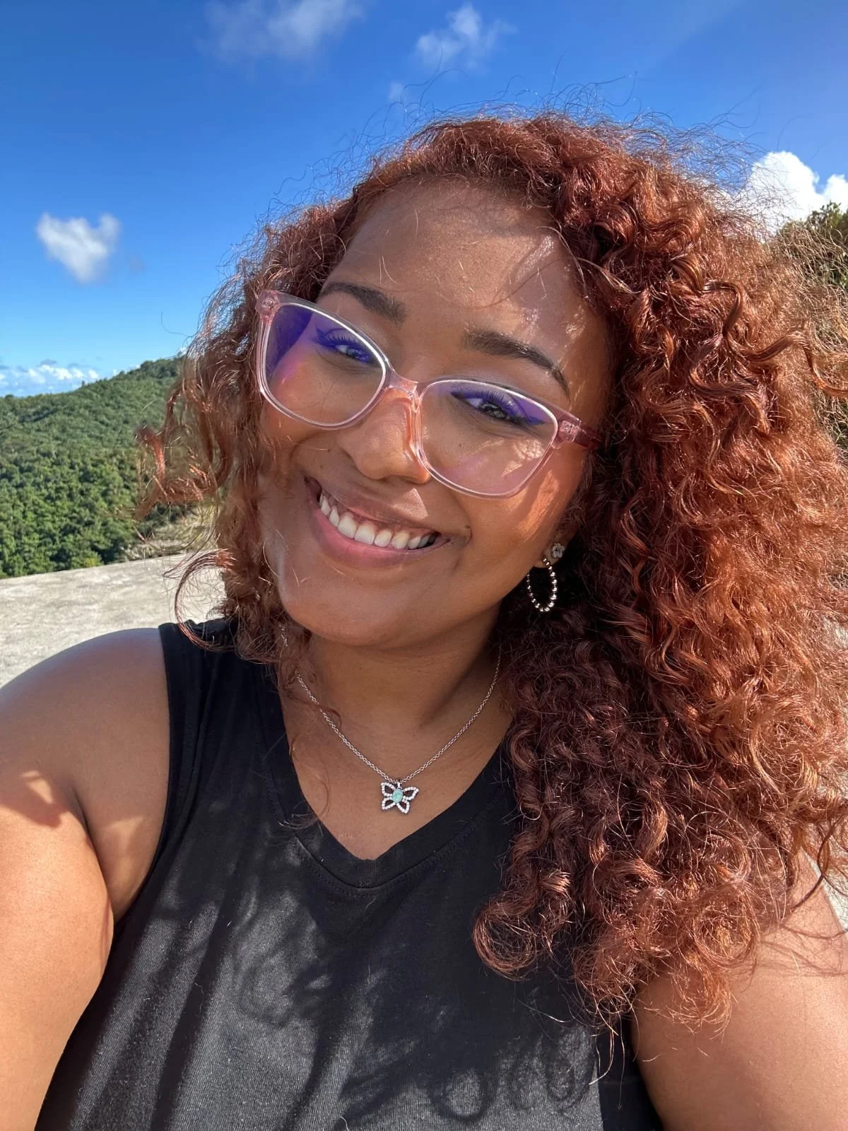 A woman with curly red hair wearing glasses, earrings, a butterfly necklace, and a black sleeveless top, smiling outdoors with a mountain and blue sky in the background.