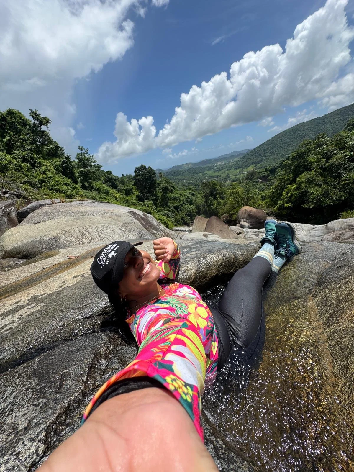 Woman lying on wet rocks in a river, smiling and taking a selfie. She is wearing a colorful floral shirt, black leggings, blue hiking shoes, a black hat, and a wristband. The scenery includes large rocks, lush green trees, mountains, and a partly cloudy sky.