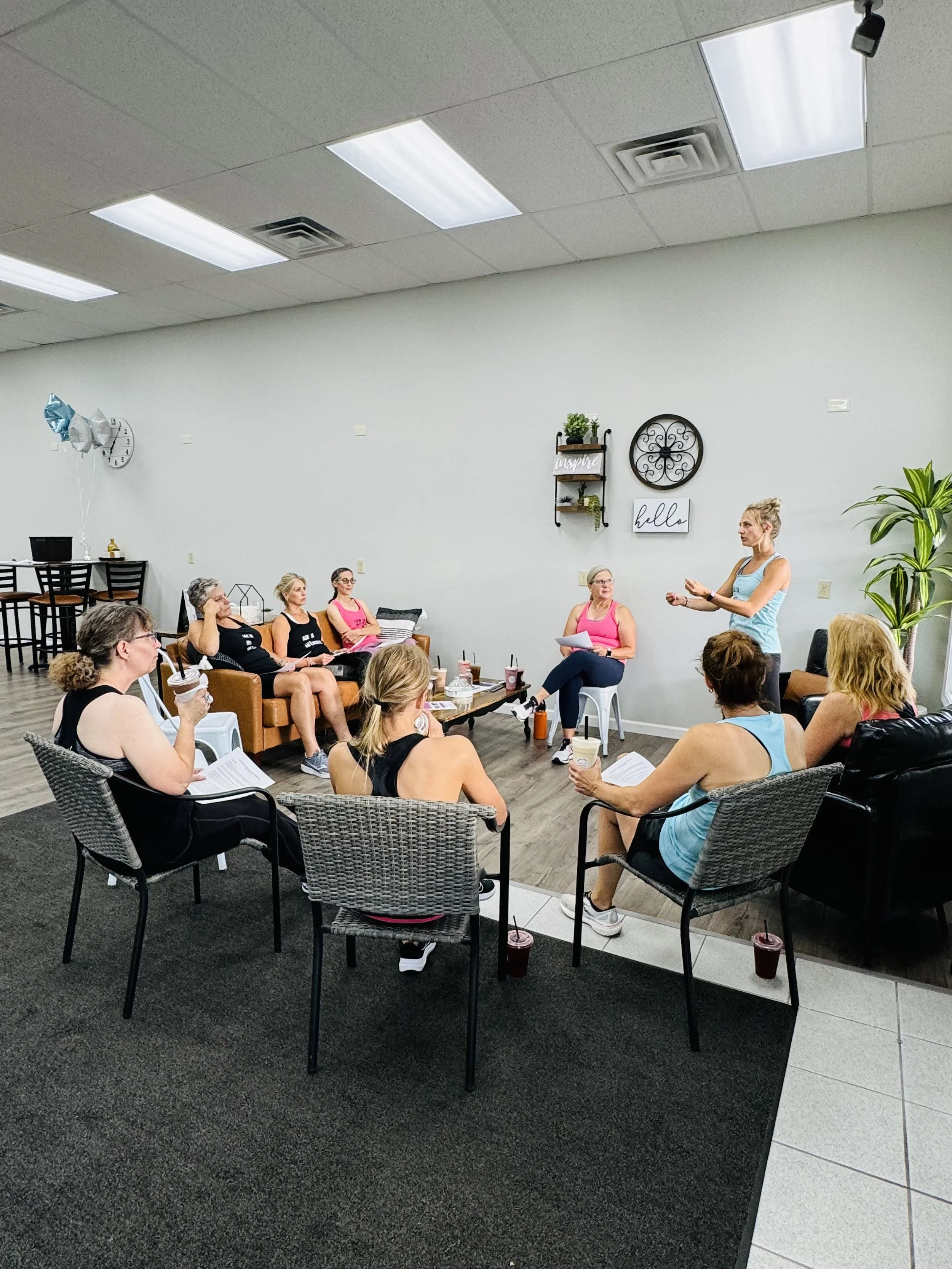 Group of women participating in a fitness class or workshop in a casual indoor setting. One woman is standing and speaking, while others sit on chairs and couches listening.