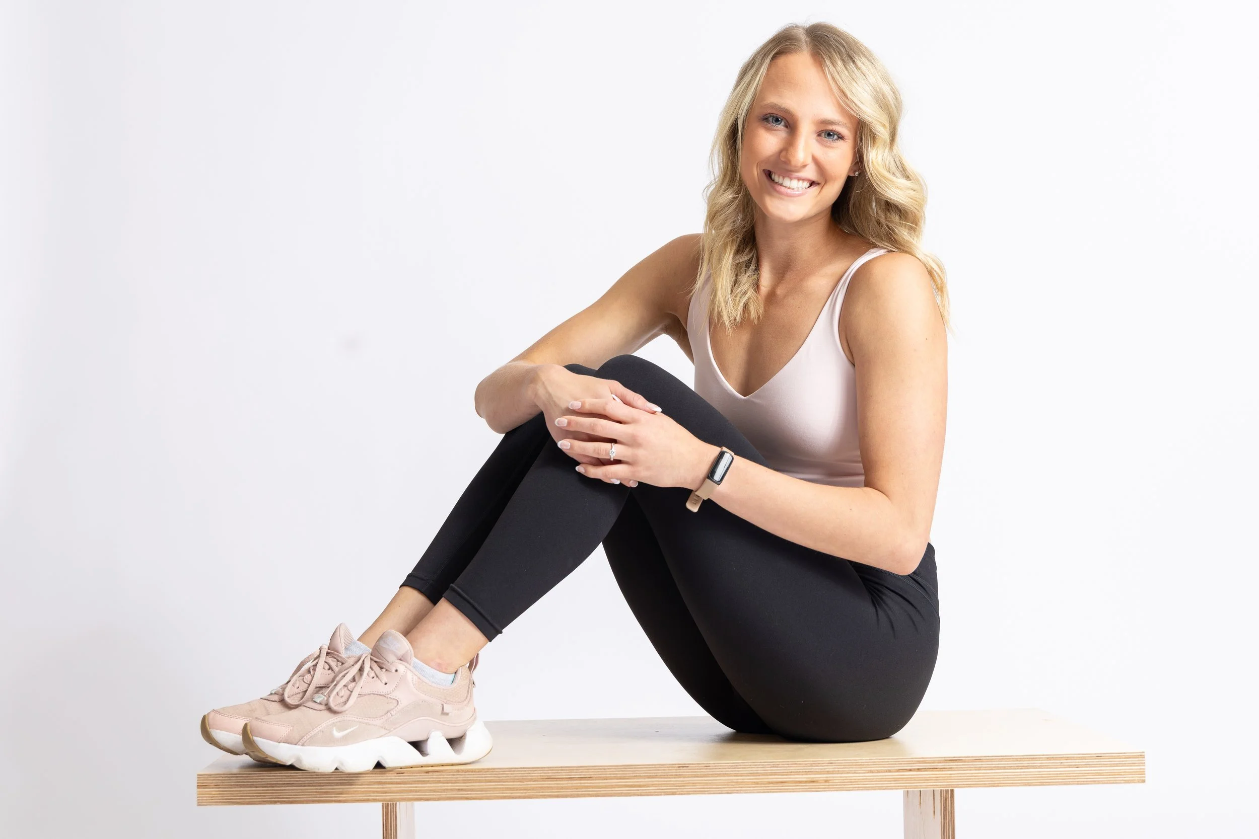 A smiling blonde woman in workout clothes sitting on a wooden bench against a white background