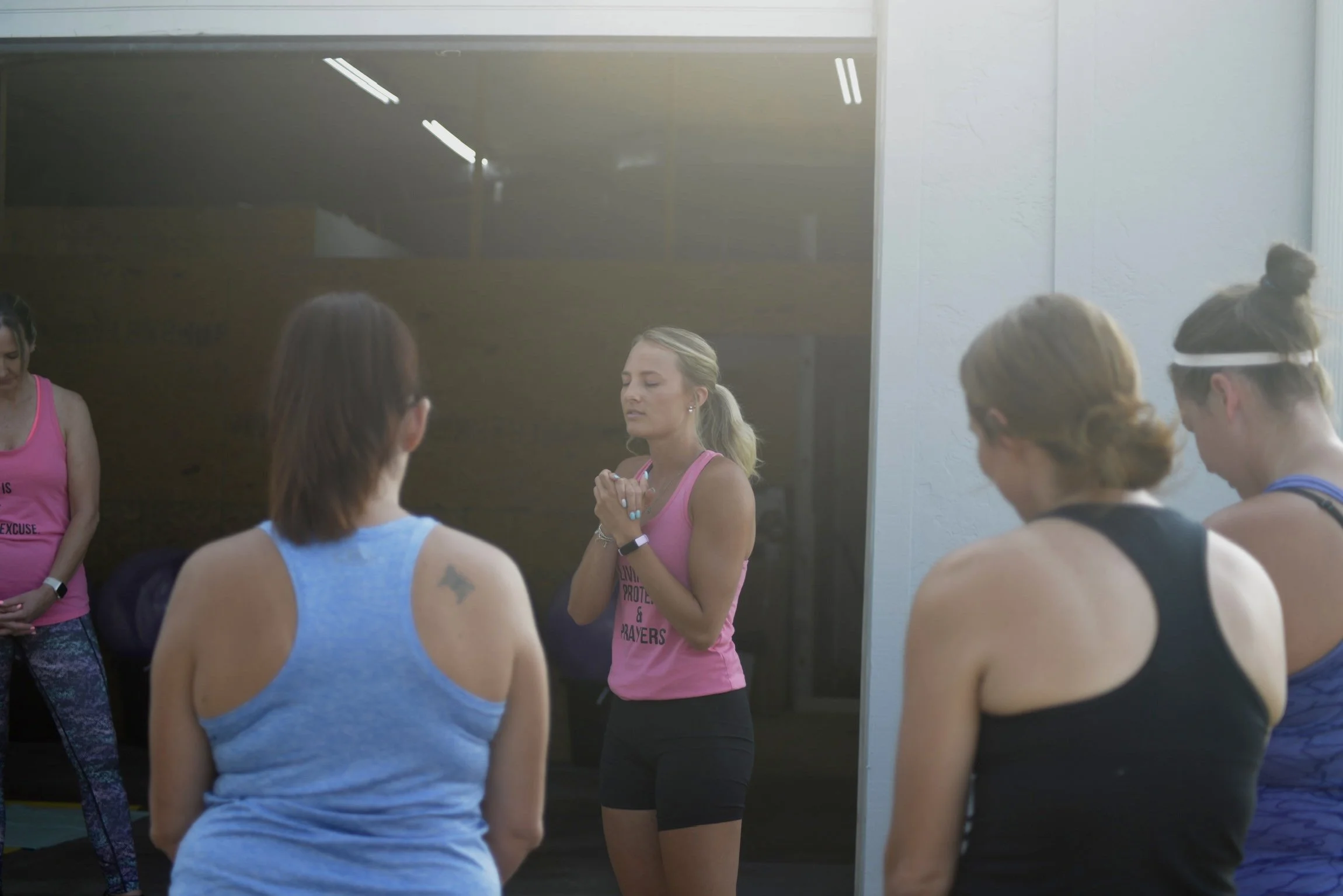 Women participating in a fitness class, standing with their heads bowed, in workout attire, in a gym or fitness studio.