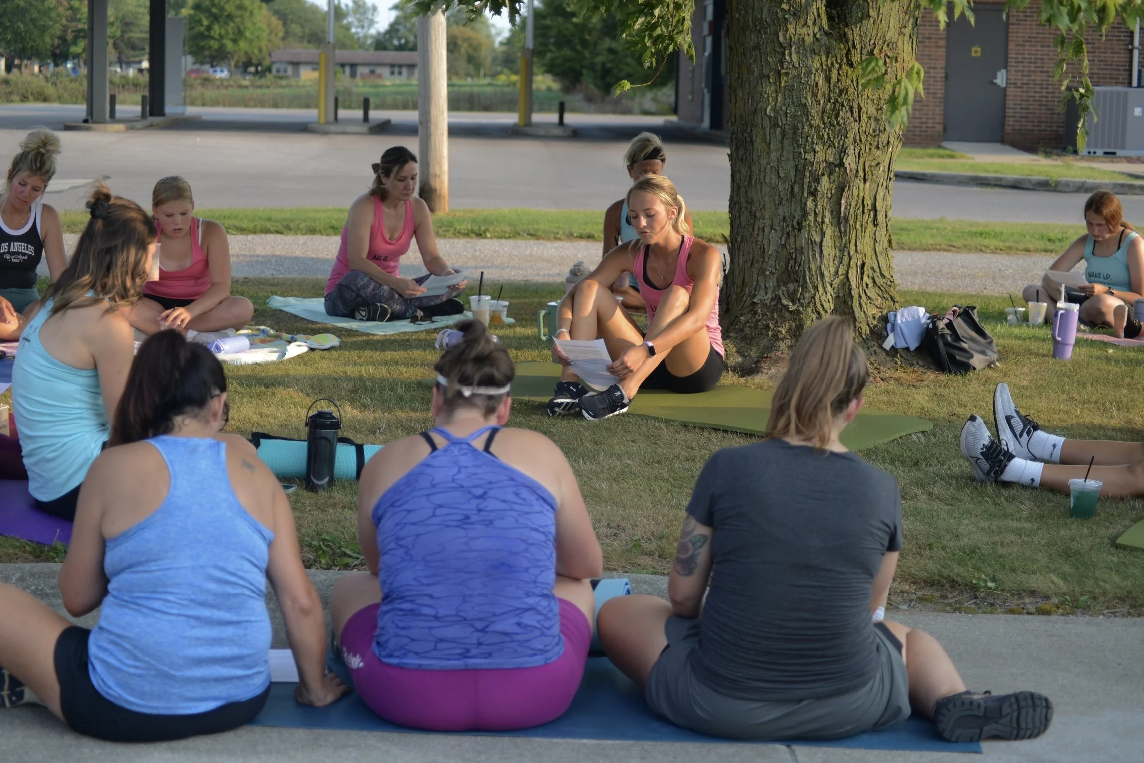 Group of women participating in an outdoor yoga class under a tree during daytime, sitting on yoga mats and wearing workout clothes.