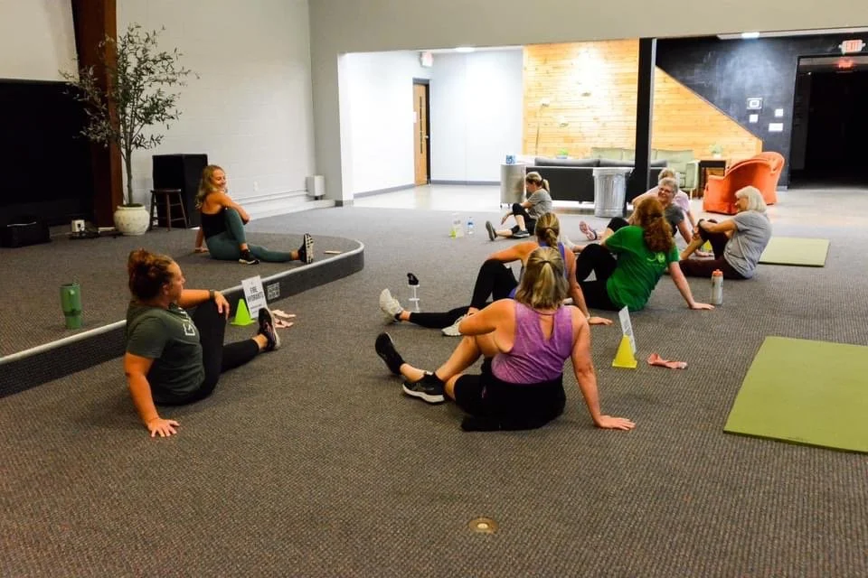 Group of women participating in a stretching or yoga class in a spacious indoor room, sitting on the floor next to yoga mats and water bottles.