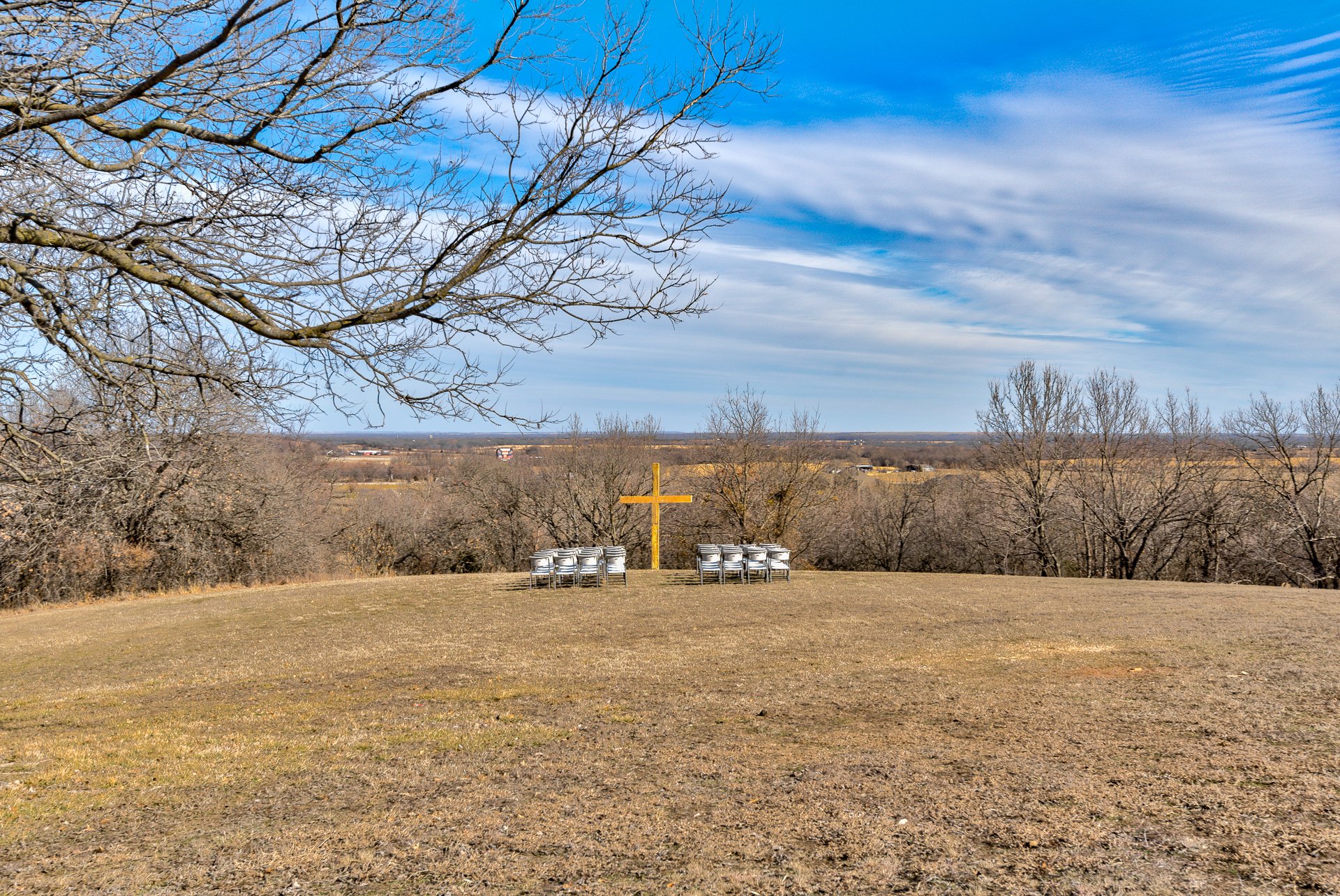 Outdoor field with a wooden cross and stacked chairs, surrounded by bare trees on a sunny day.