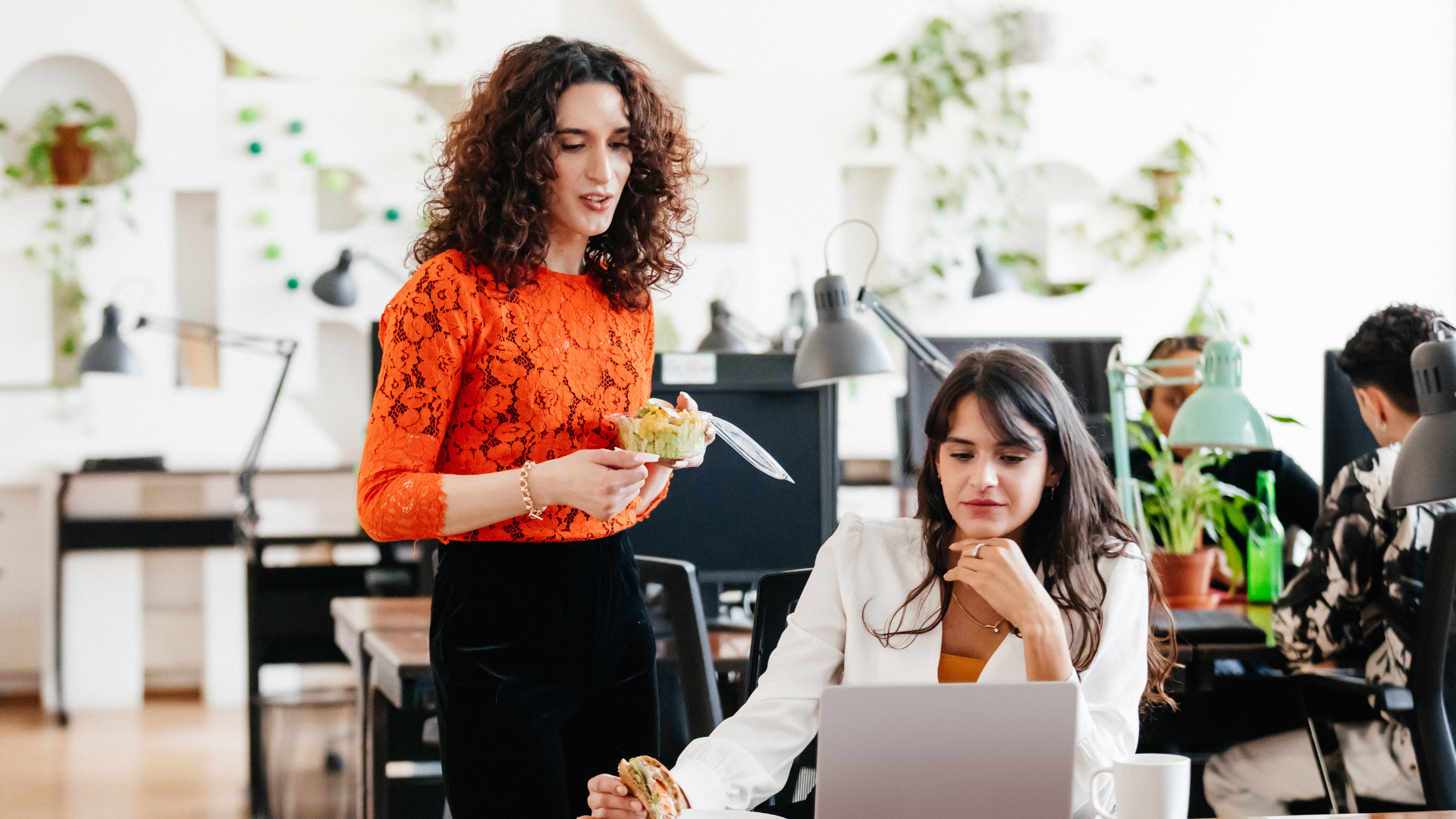 Two women in an office, one standing with a salad and the other sitting with a sandwich and a laptop, surrounded by office equipment and plants.