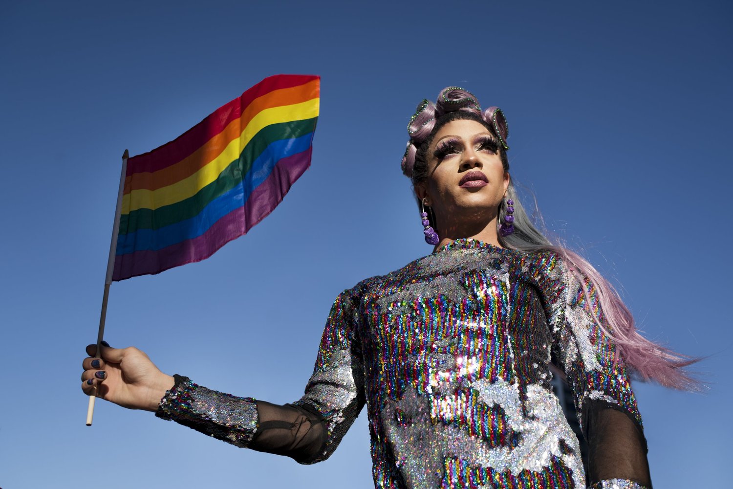 A person in a sparkling outfit holding a rainbow flag beneath a blue sky.