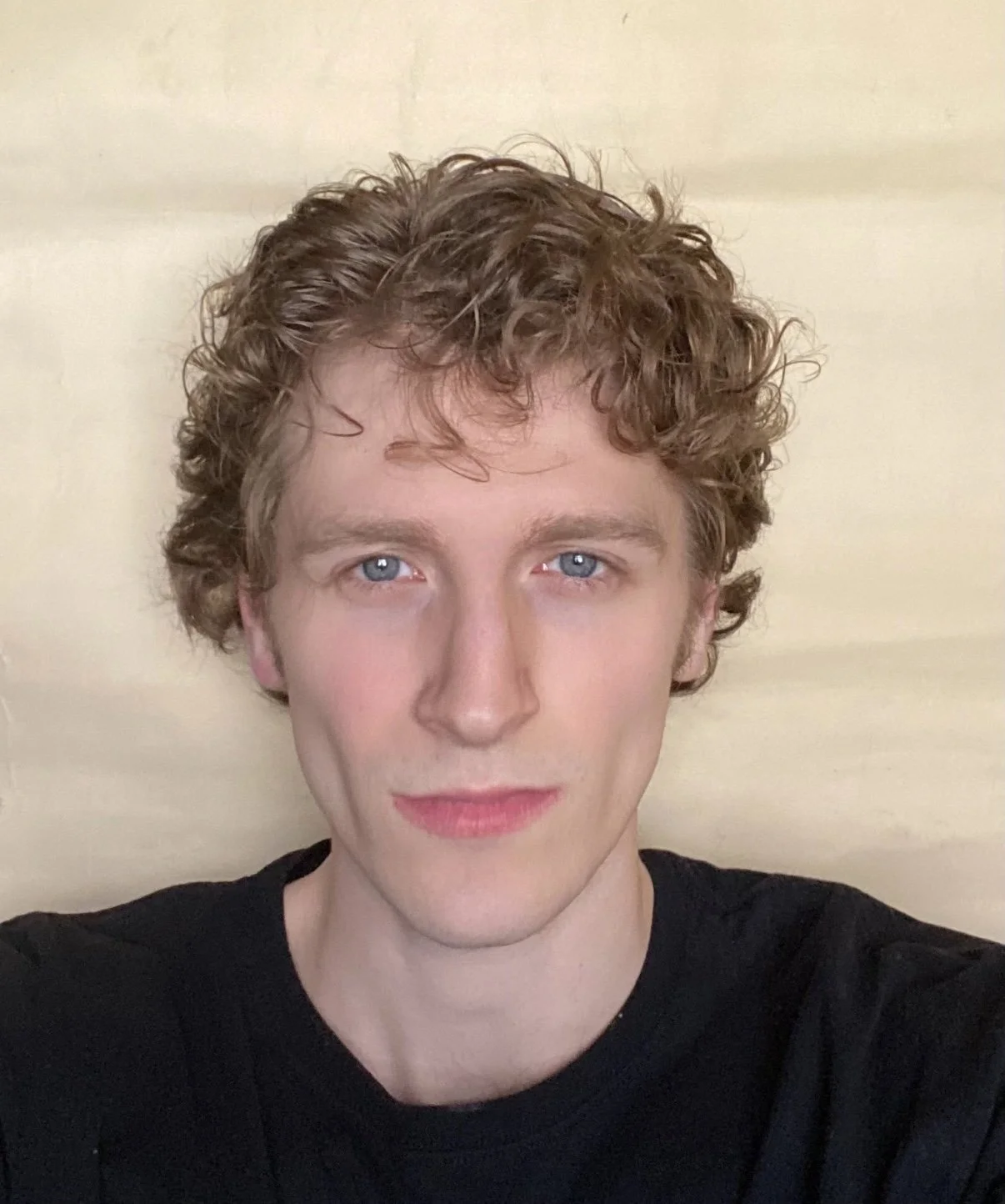 A young man with curly light brown hair and fair skin taking a selfie outdoors with lush green foliage and small white flowers in the background.