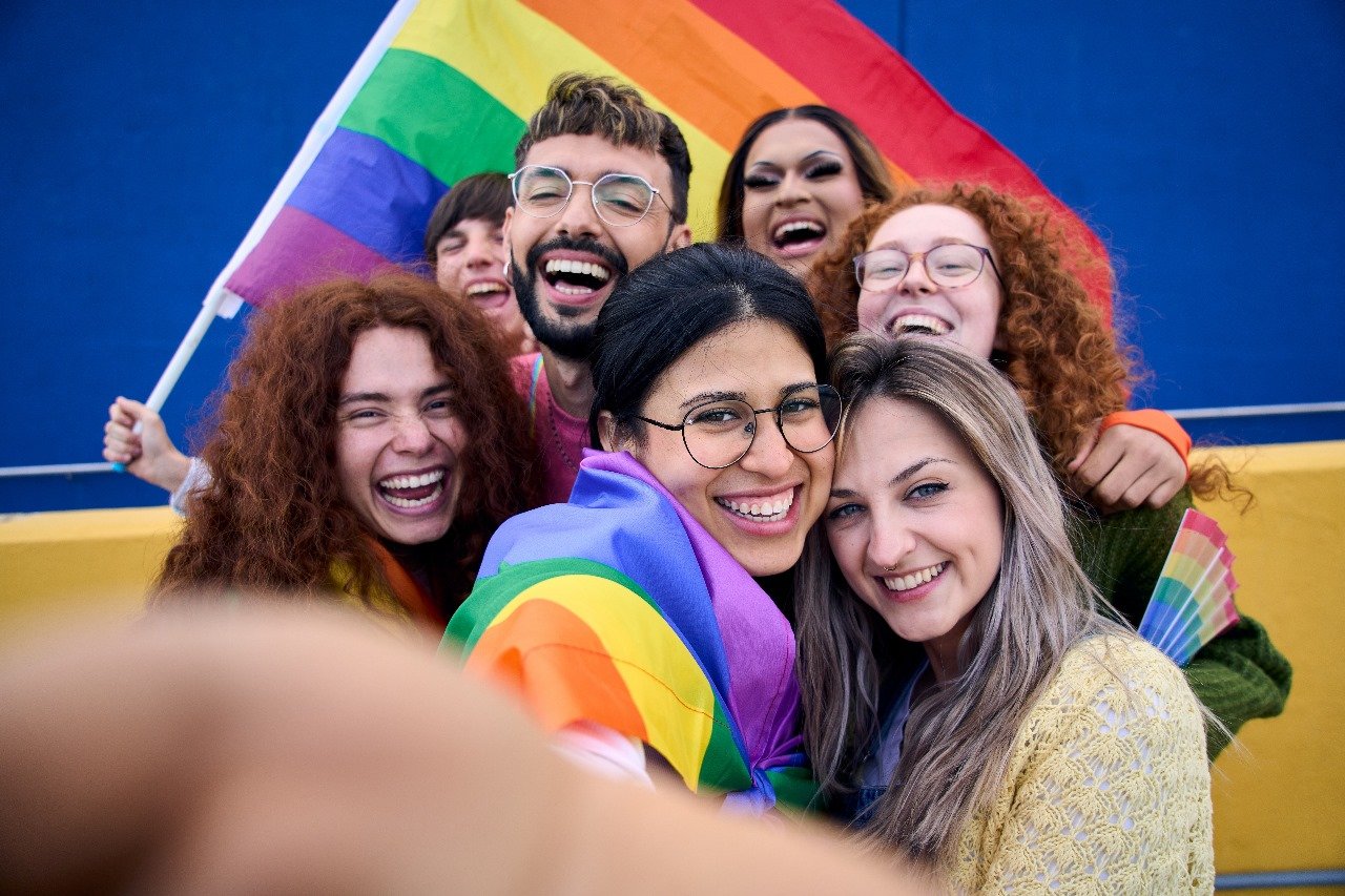 A group of people smiling together in front of a rainbow flag.