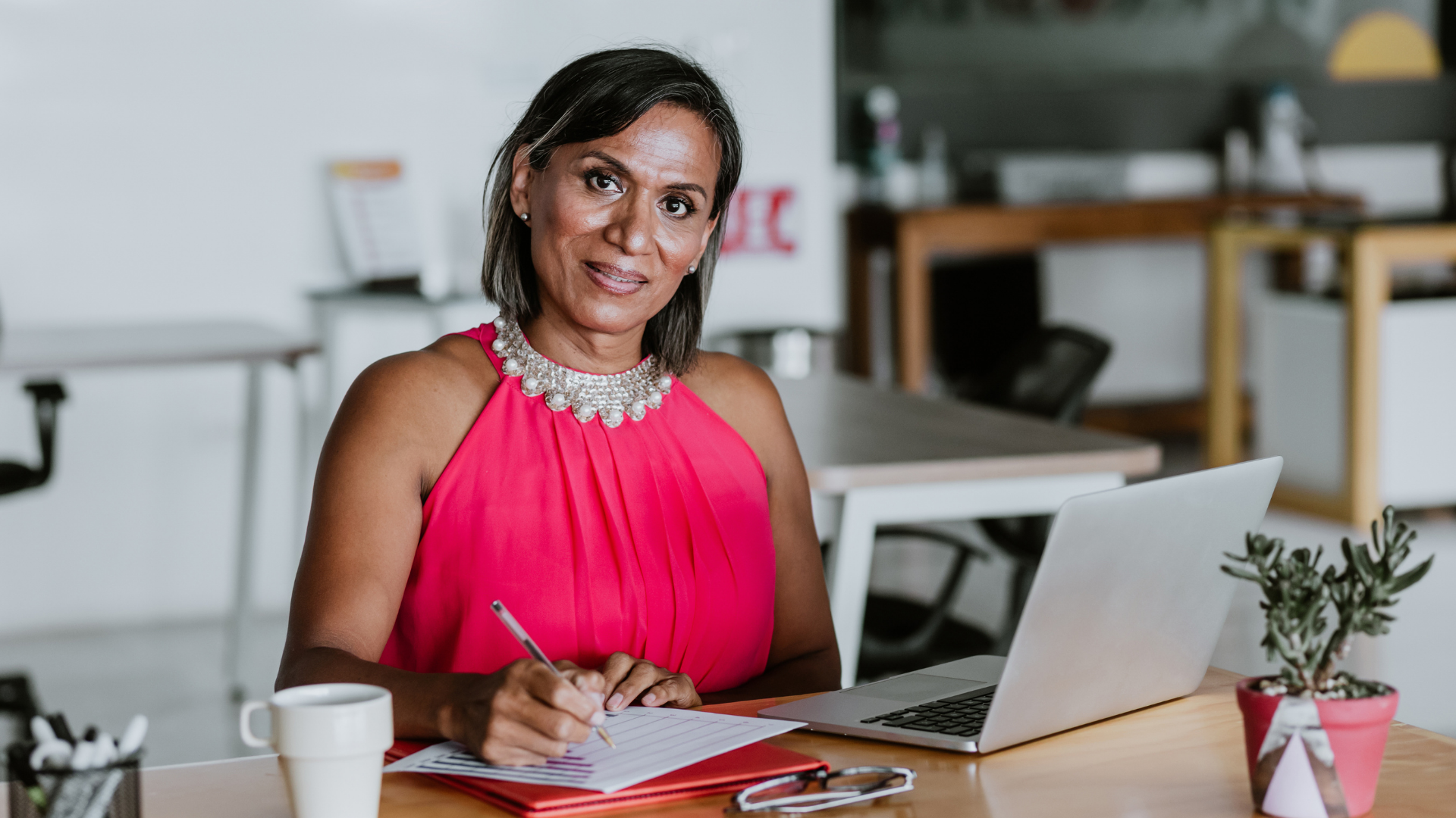 A woman in a pink dress sitting at a desk with a laptop, notebook, pen, and a small potted plant.