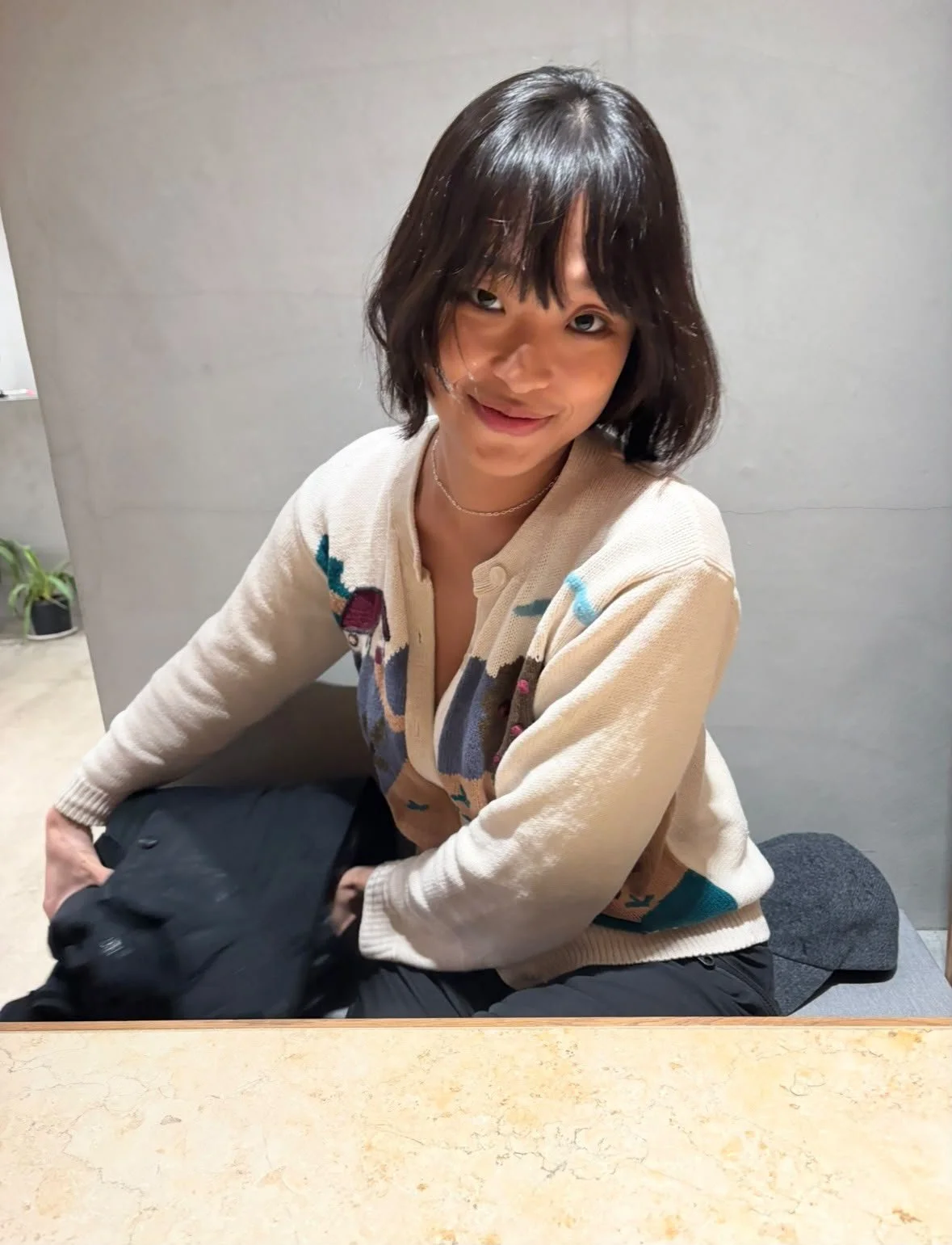 A woman with short dark hair and bangs, wearing a beige sweater with a colorful pattern, sitting at a table with a black dog on her lap, smiling at the camera.