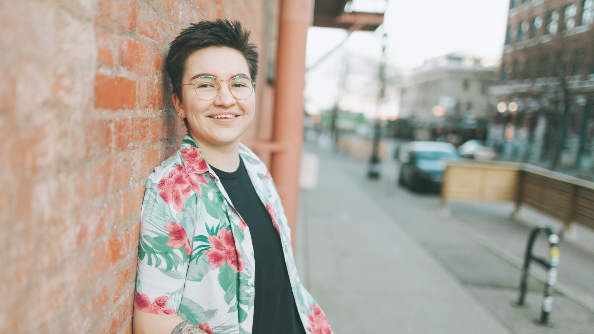 A young person with short dark hair, glasses, and a floral shirt smiling while leaning against a brick wall on a city sidewalk.