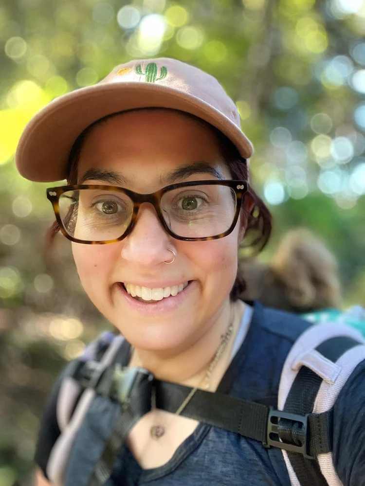 Madrone Phoenix smiling outdoors, wearing glasses and a cap.