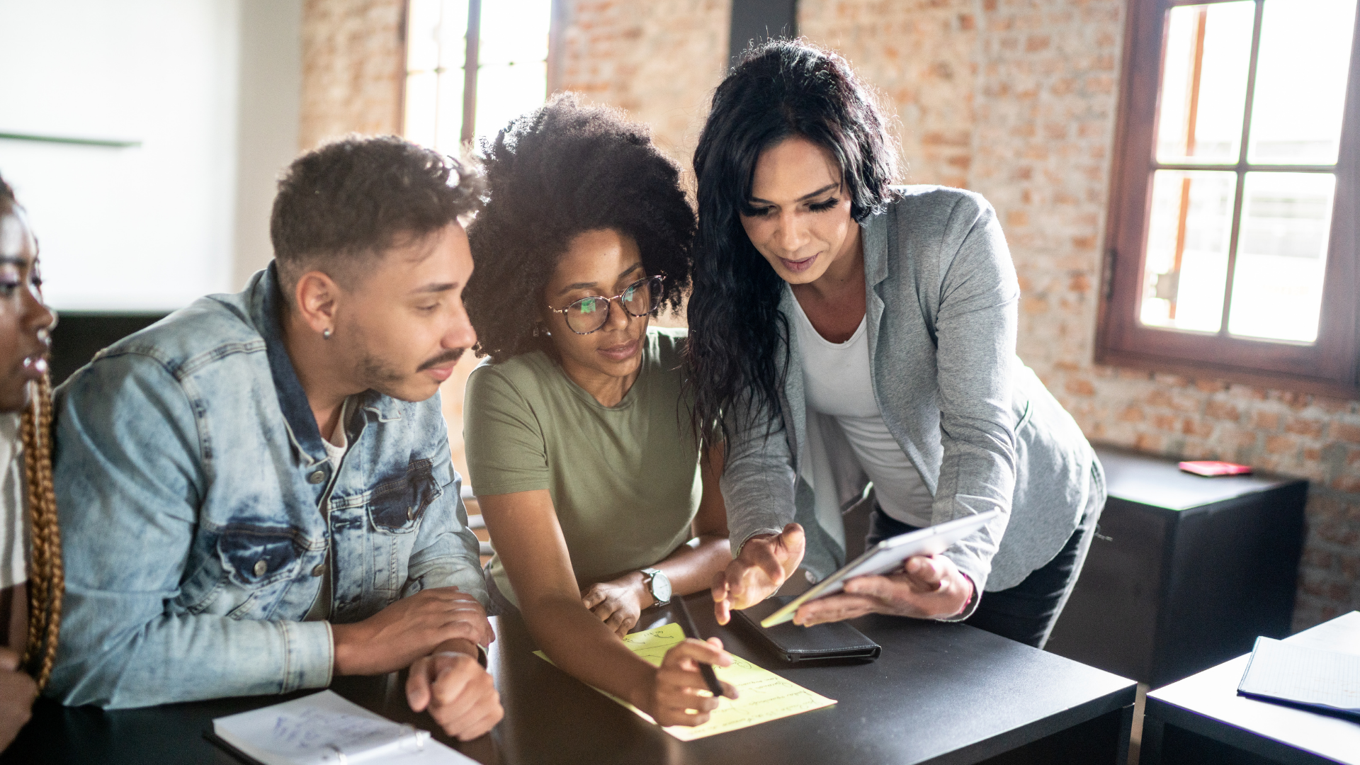 Four diverse people in a meeting, two women and two men, gathered around a table, focused on a tablet held by a woman with dark wavy hair, in a room with brick walls and large windows.