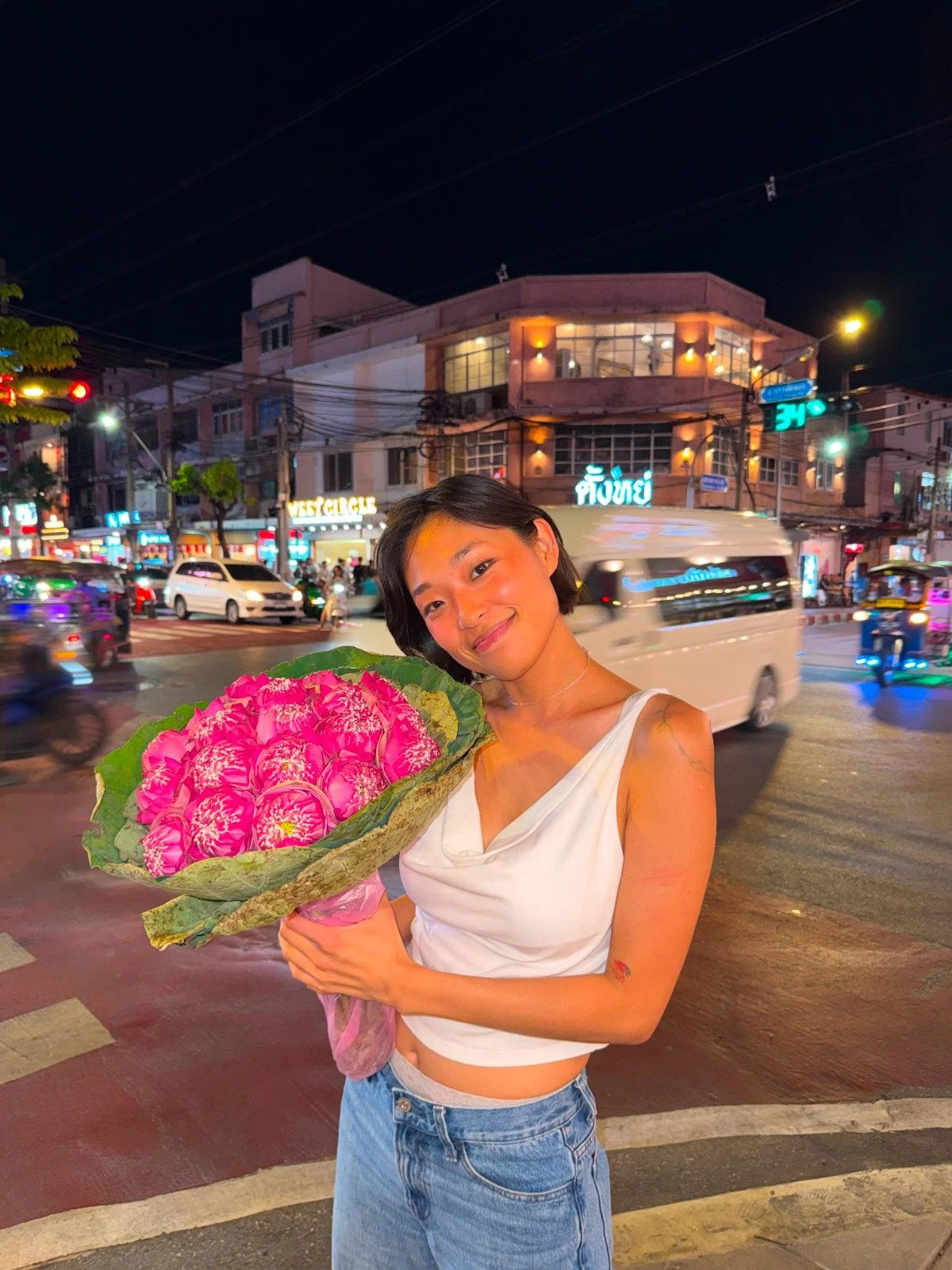 A woman smiling and holding a large bouquet of pink flowers on a busy city street at night with cars and neon signs in the background.