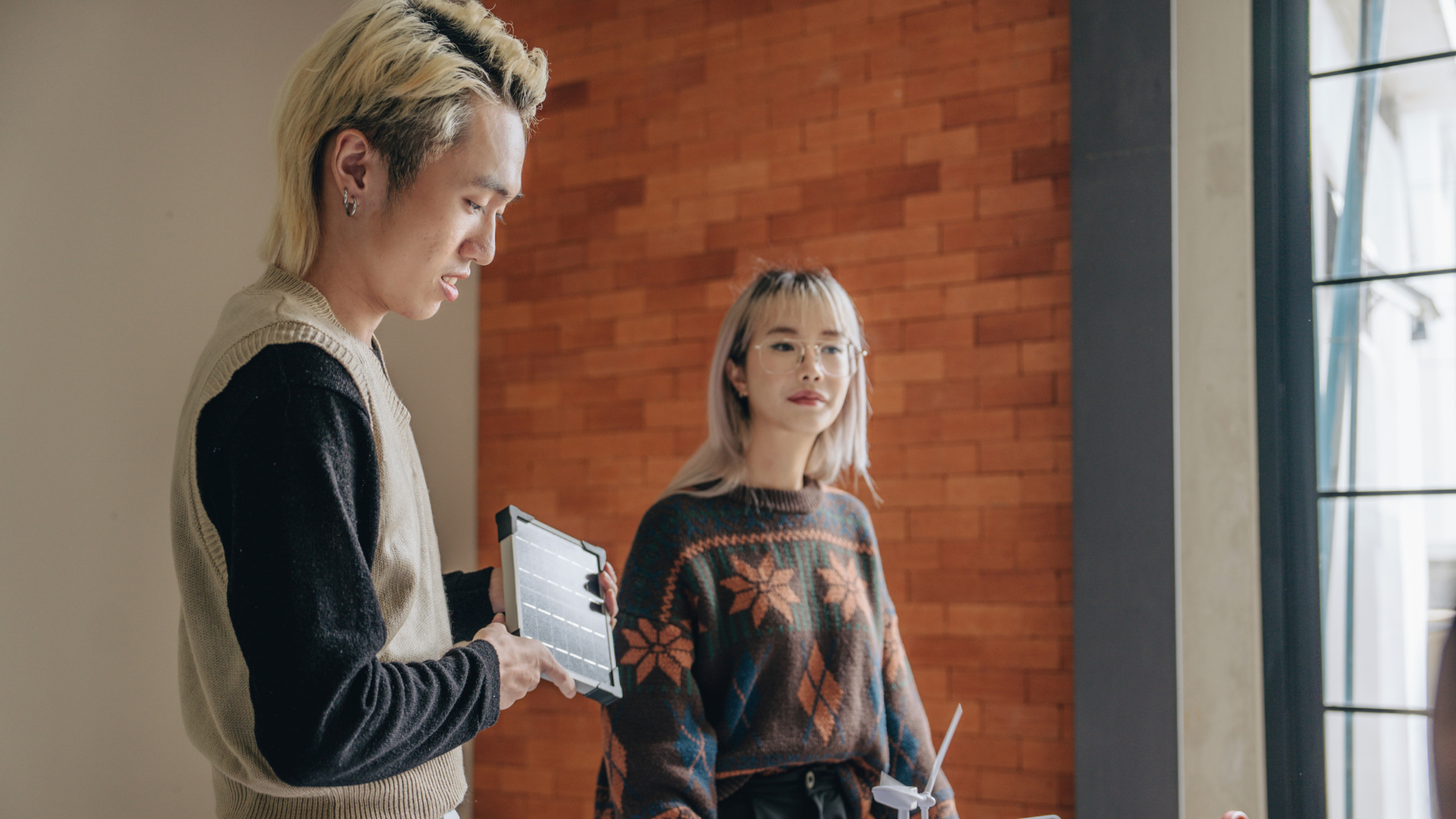 Two women are inside a room with a brick wall and large window. One woman is holding a tablet and looking at the other woman, who is wearing glasses and holding a tablet. The second woman is looking at the first woman.