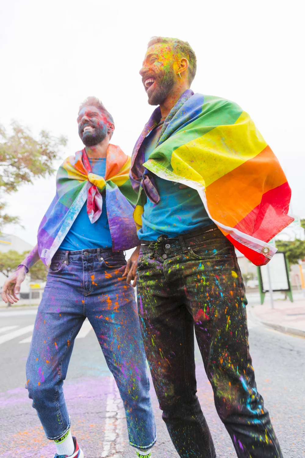 Two people laughing outdoors, wearing rainbow flags and covered in colorful powder.
