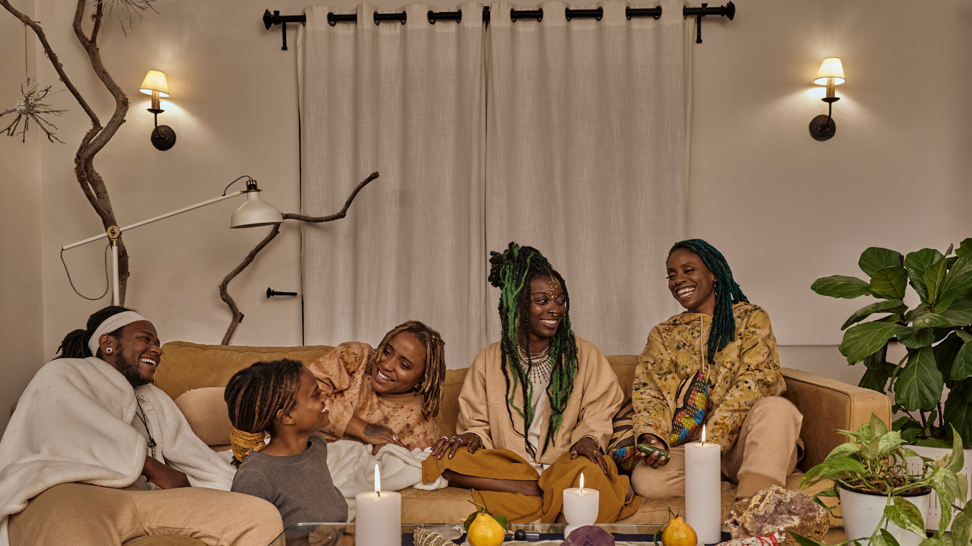 A group of six people, four women and two men, sitting on a beige couch in a cozy living room, smiling and enjoying each other's company. The room features warm lighting, green plants, and candles, creating a relaxed and joyful atmosphere.