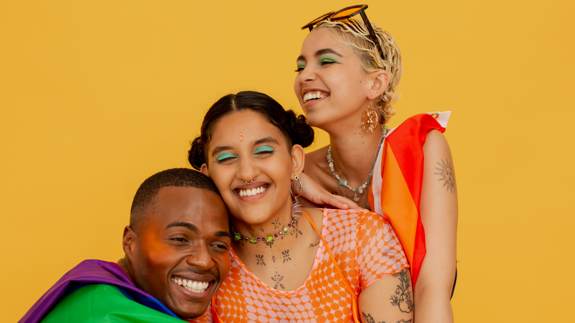 Three happy people with diverse styles and colorful makeup, embracing and smiling against a yellow background.