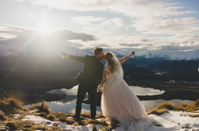A newlywed couple eloping in New Zealand. They stand on a hilltop with snow, holding hands and leaning in for a kiss, with a scenic mountain and lake landscape in the background during sunset.