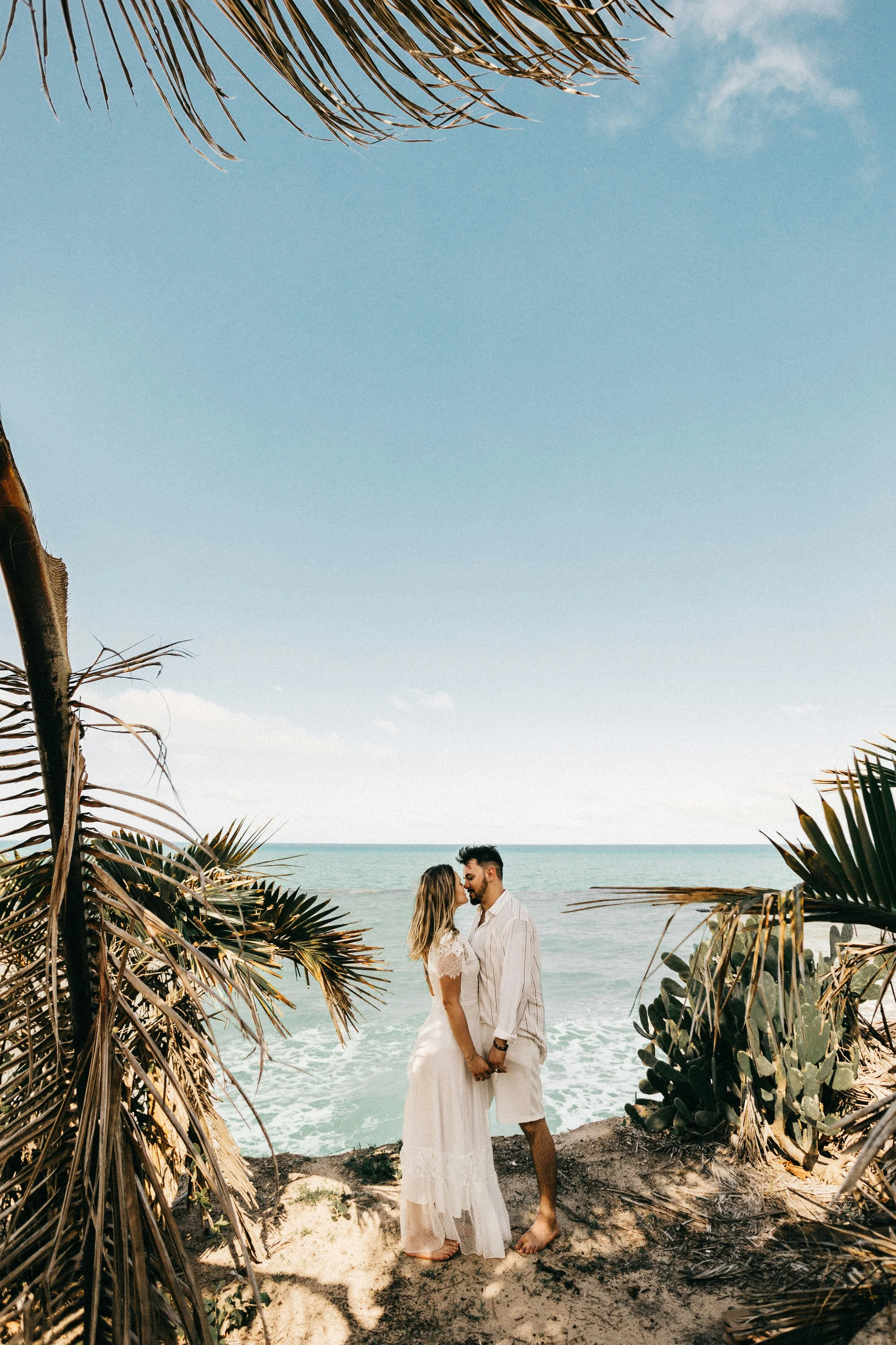 A couple dressed in white stand close together on a sandy beach, holding hands and touching foreheads, surrounded by tropical plants, with the ocean and blue sky in the background.
