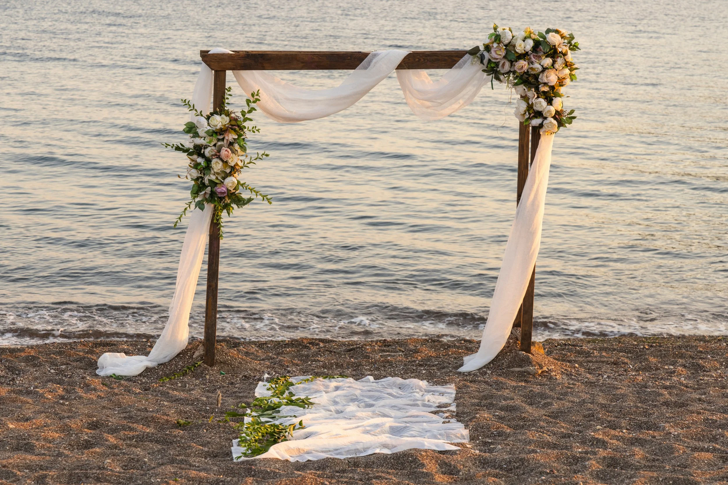Wedding arch decorated with flowers and fabric on a beach with a fabric aisle and greenery.