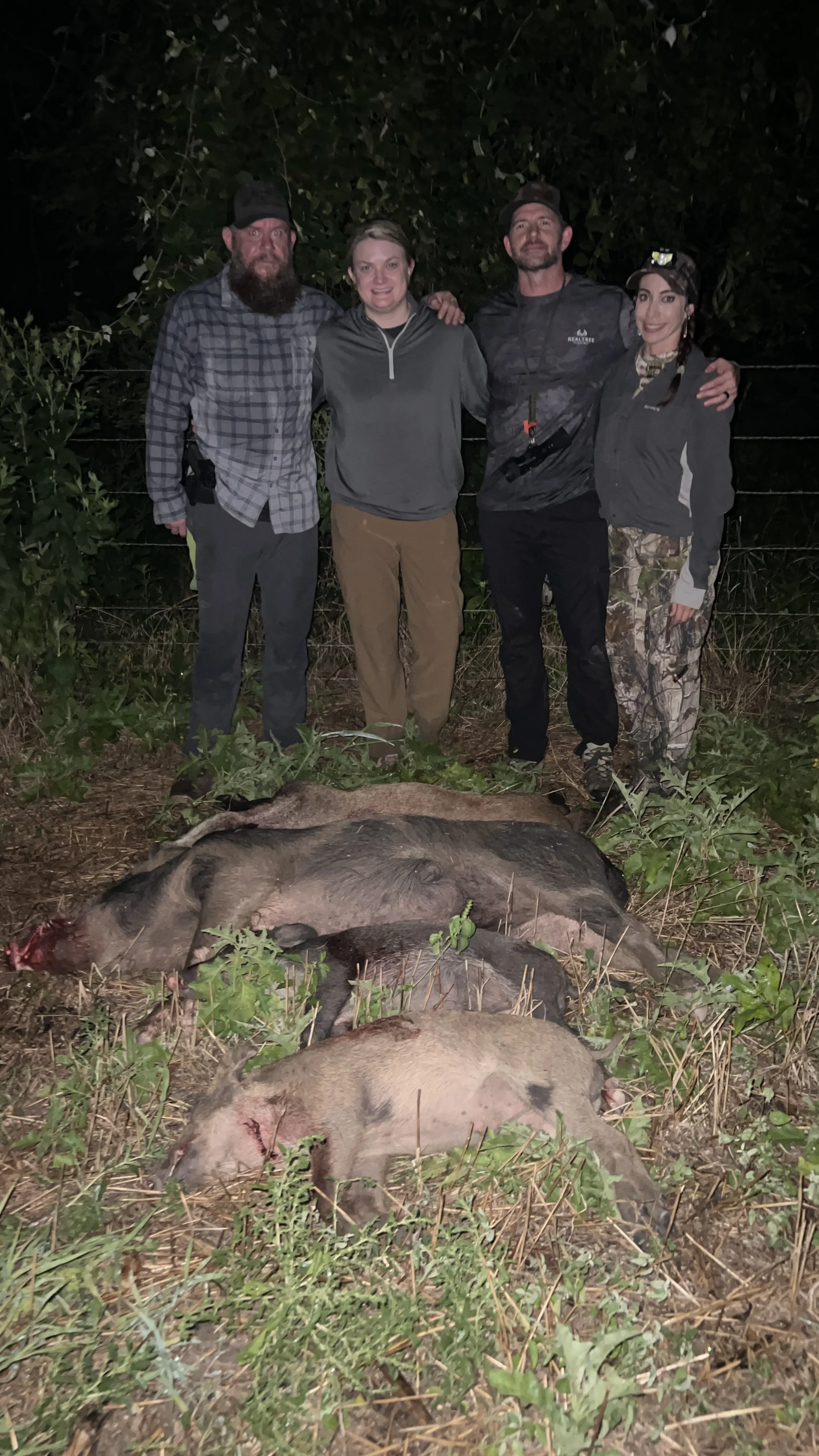 Group of four people standing outdoors at night, posing with a hunted wild pig on the ground in front of them.